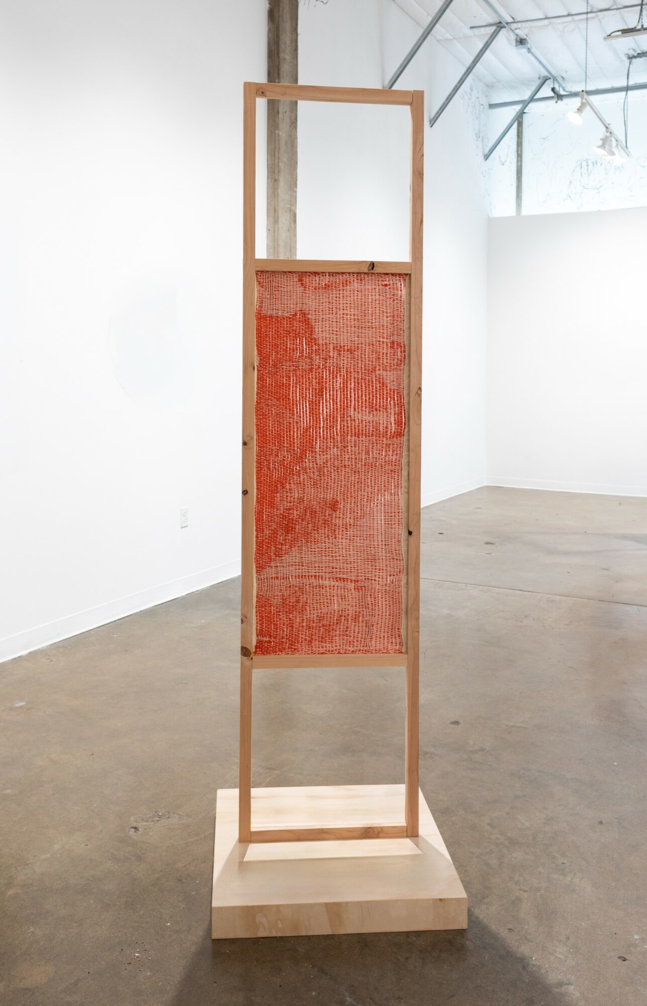 Tall wooden frame with a red textured panel, standing on a base in an art gallery with white walls and a concrete floor.