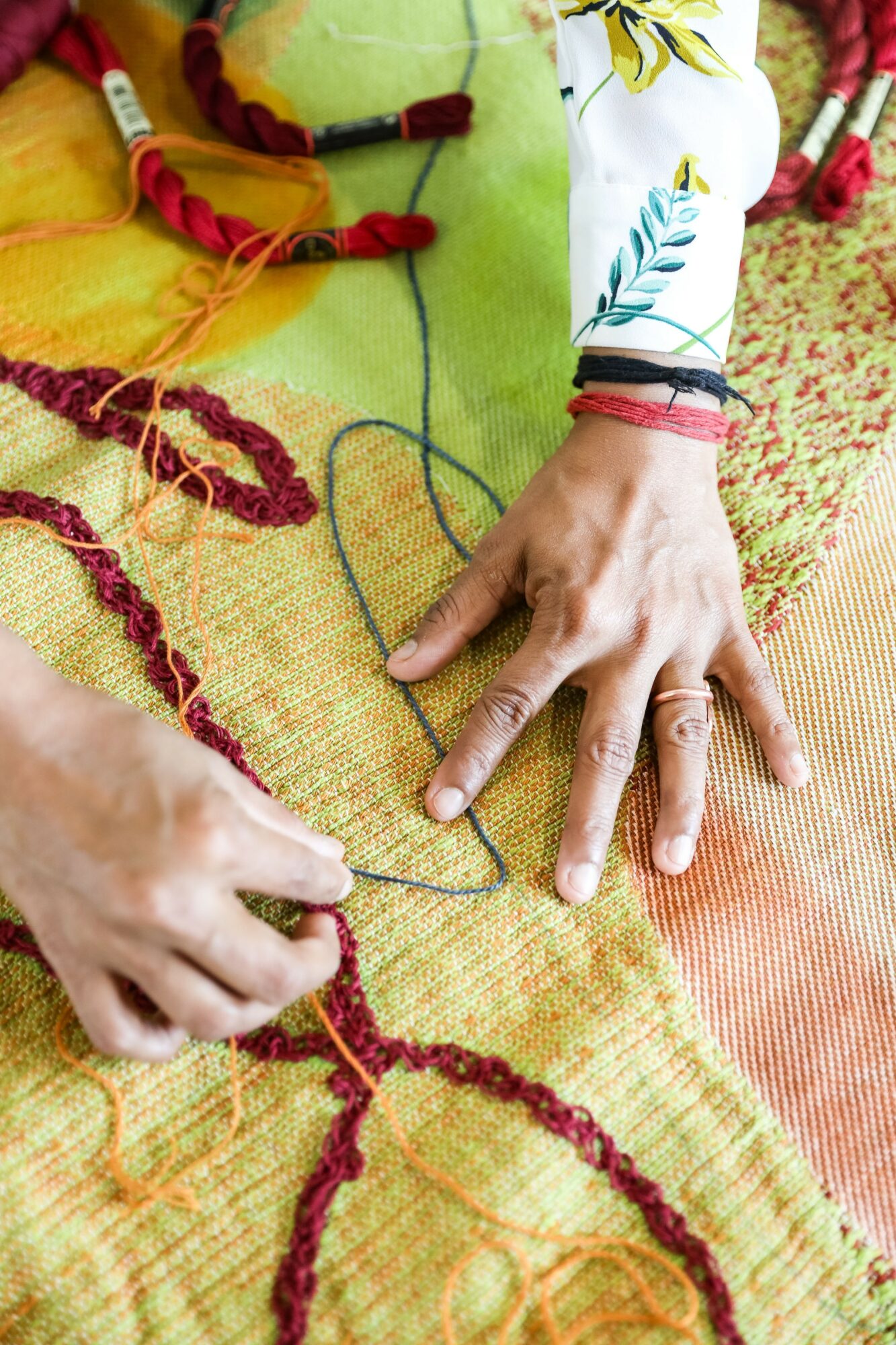 Person's hand holding a needle and thread, working on colorful fabric with red and yellow patterns, wearing a white sleeve with embroidery.