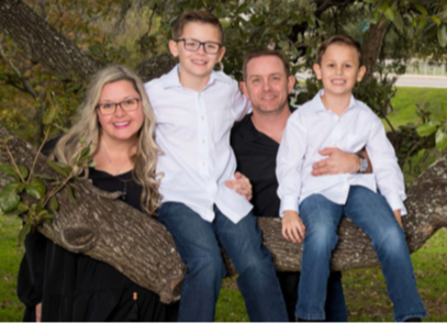 Family of four sitting on a tree branch outdoors, smiling, with trees and grass in background.