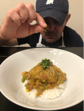 Person holding a small green item above a plate of yellow curry with rice, garnished with herbs.
