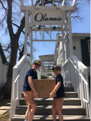 Two children holding a large box on stairs outside a house, with a sign reading 'Olamaie' above them.