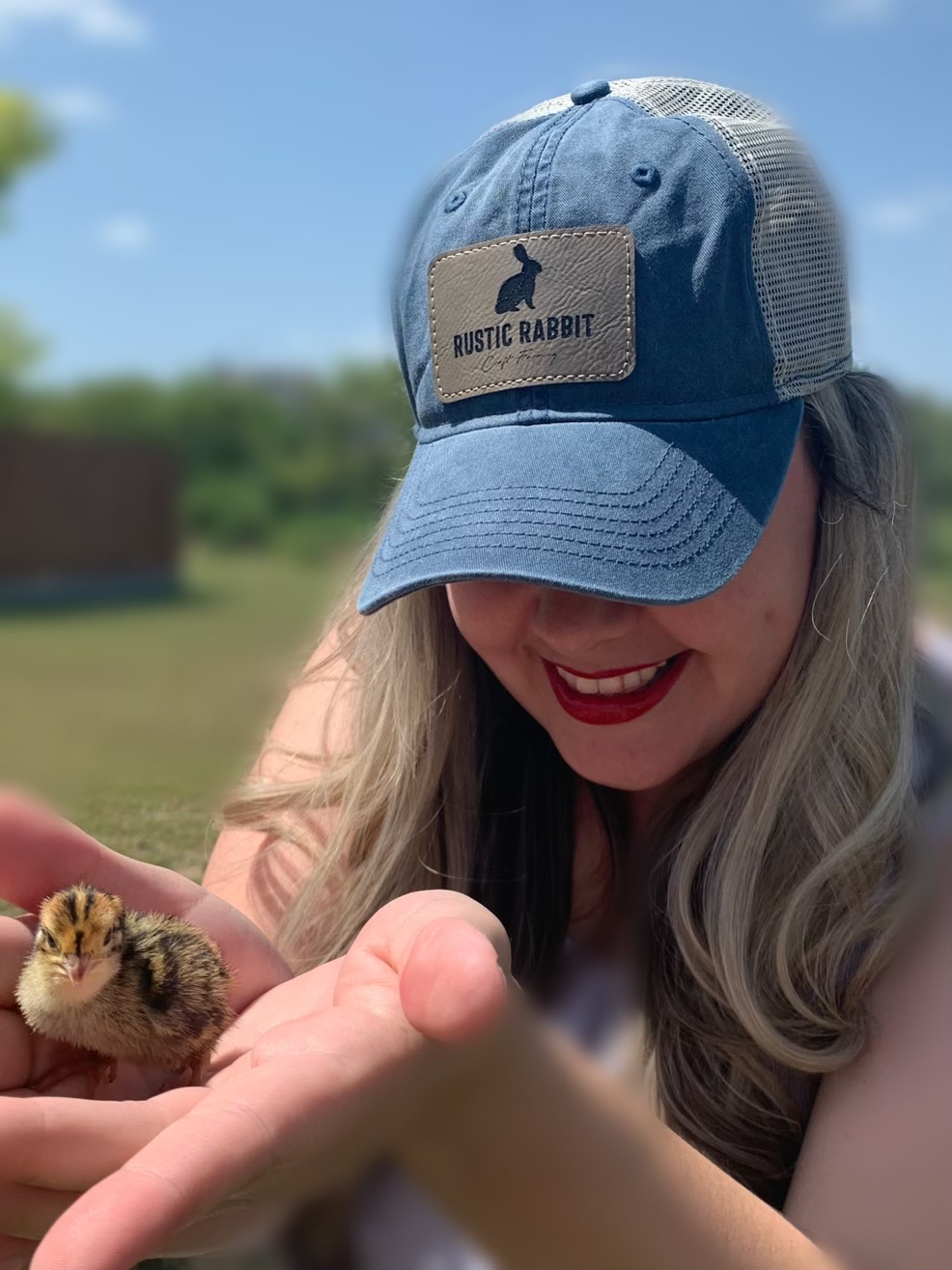 Woman smiling and holding a small chick outdoors, wearing a blue cap with a patch that says 'Music Rabbit'.