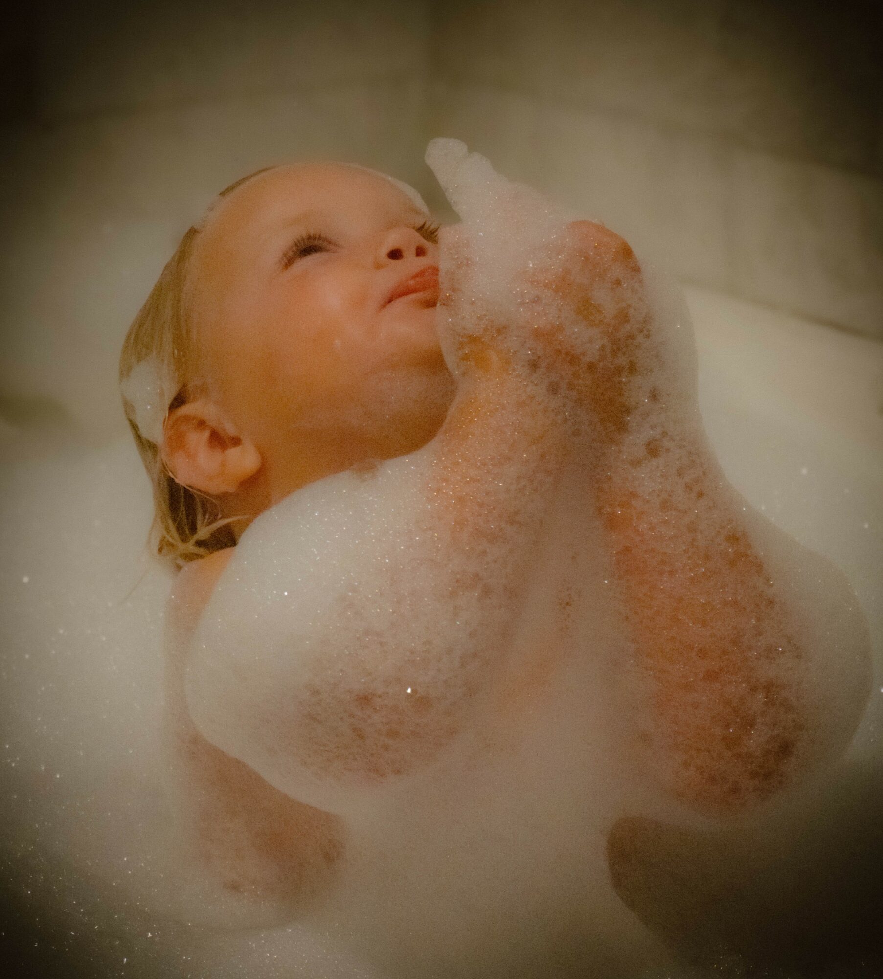 Child with blonde hair enjoying a bubble bath, surrounded by foam, looking up with a relaxed expression.