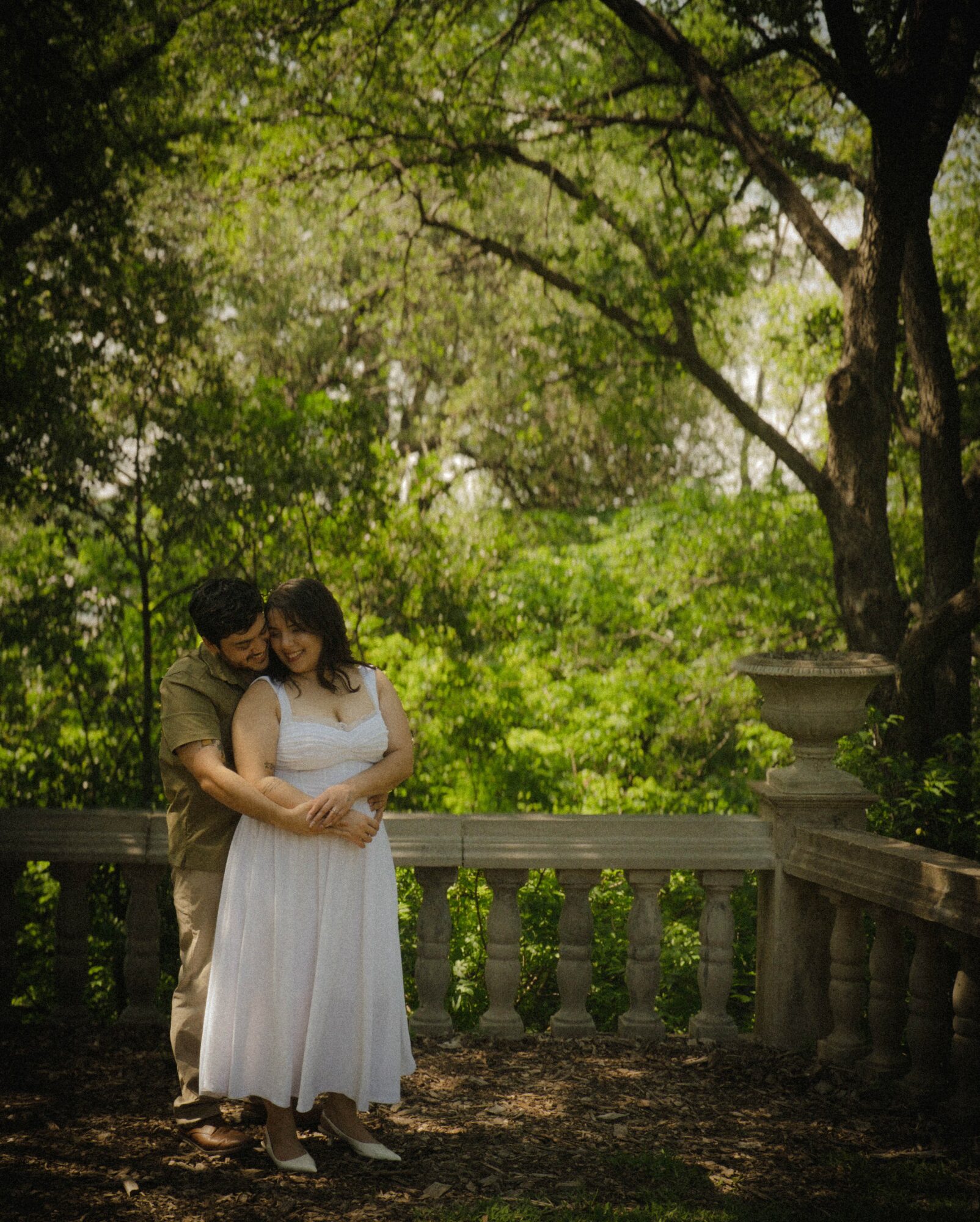 A couple stands close together on a balcony surrounded by lush green trees, with sunlight filtering through the leaves.
