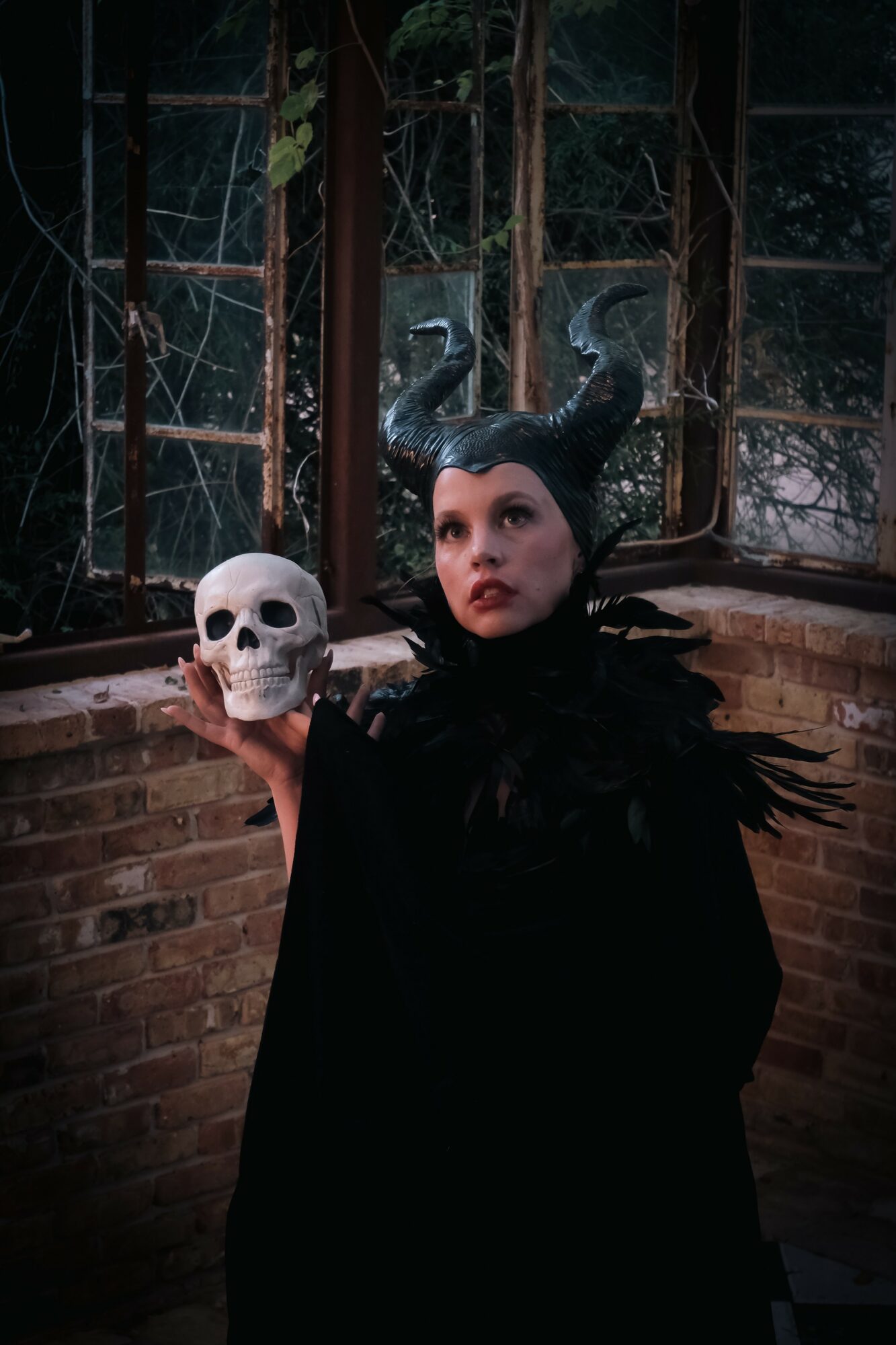 Person dressed as a witch with horns holding a skull, standing in front of broken windows and brick wall.