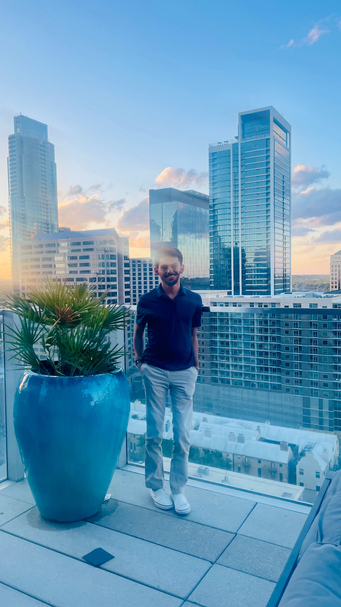 Man standing on rooftop with city skyline and tall buildings in background, large blue plant pot nearby, sunset sky.