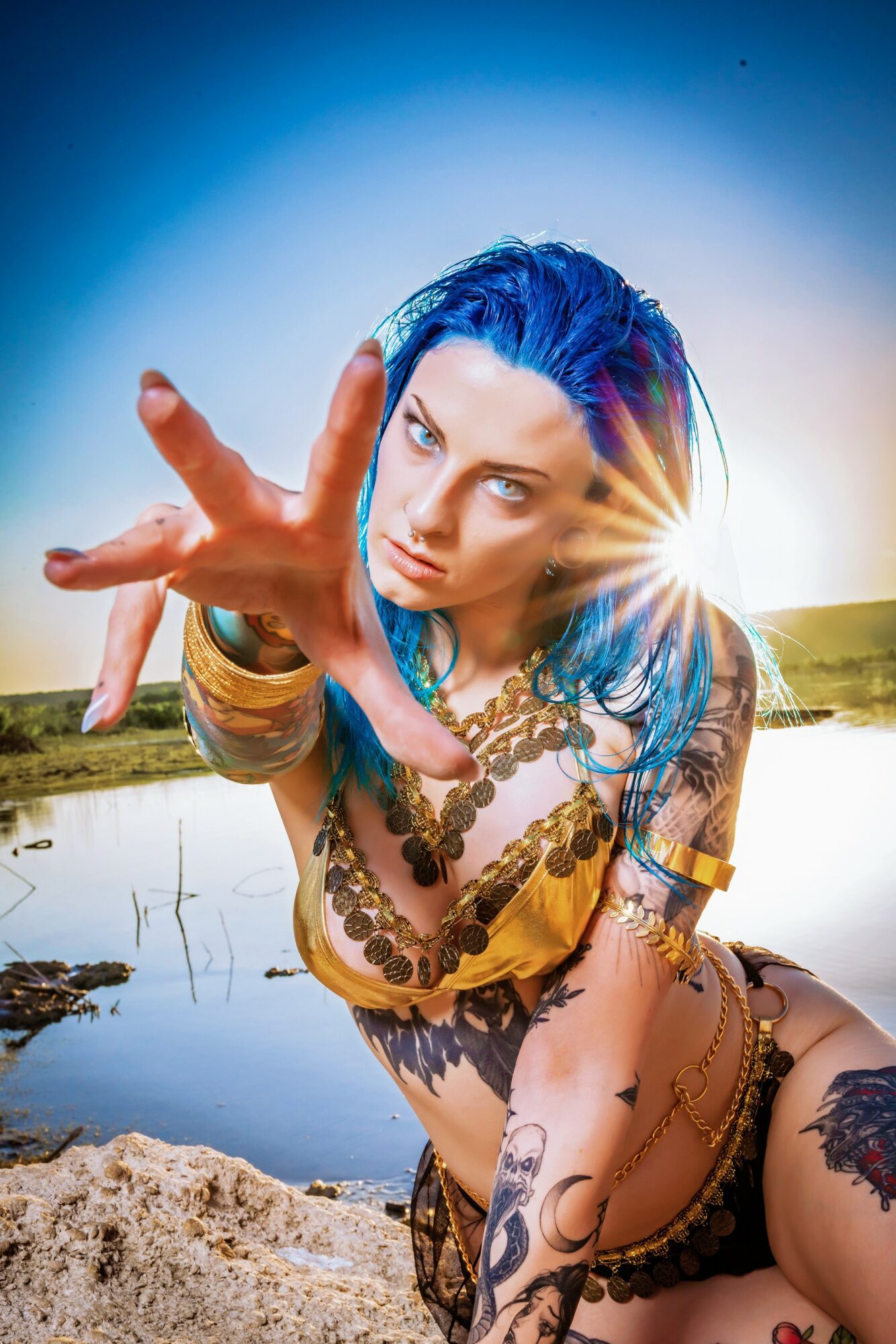 Woman with blue hair reaching toward the camera outdoors near water, sunlight shining behind her, wearing jewelry and patterned clothing.