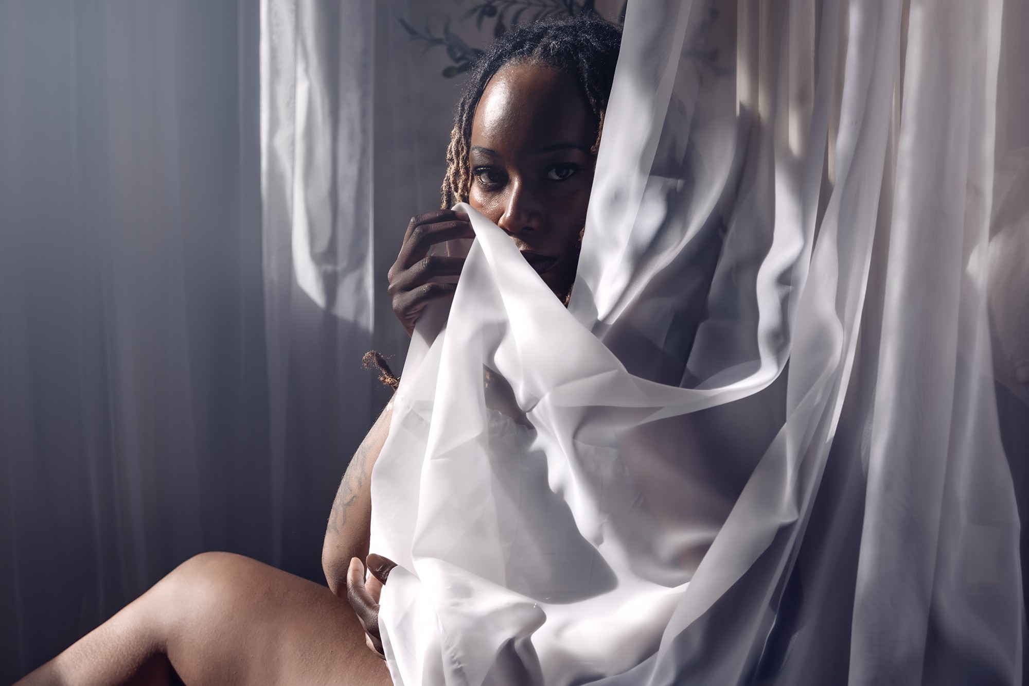 Person partially hidden behind white curtains, holding fabric, with dark hair and eyes visible, in a dimly lit room.
