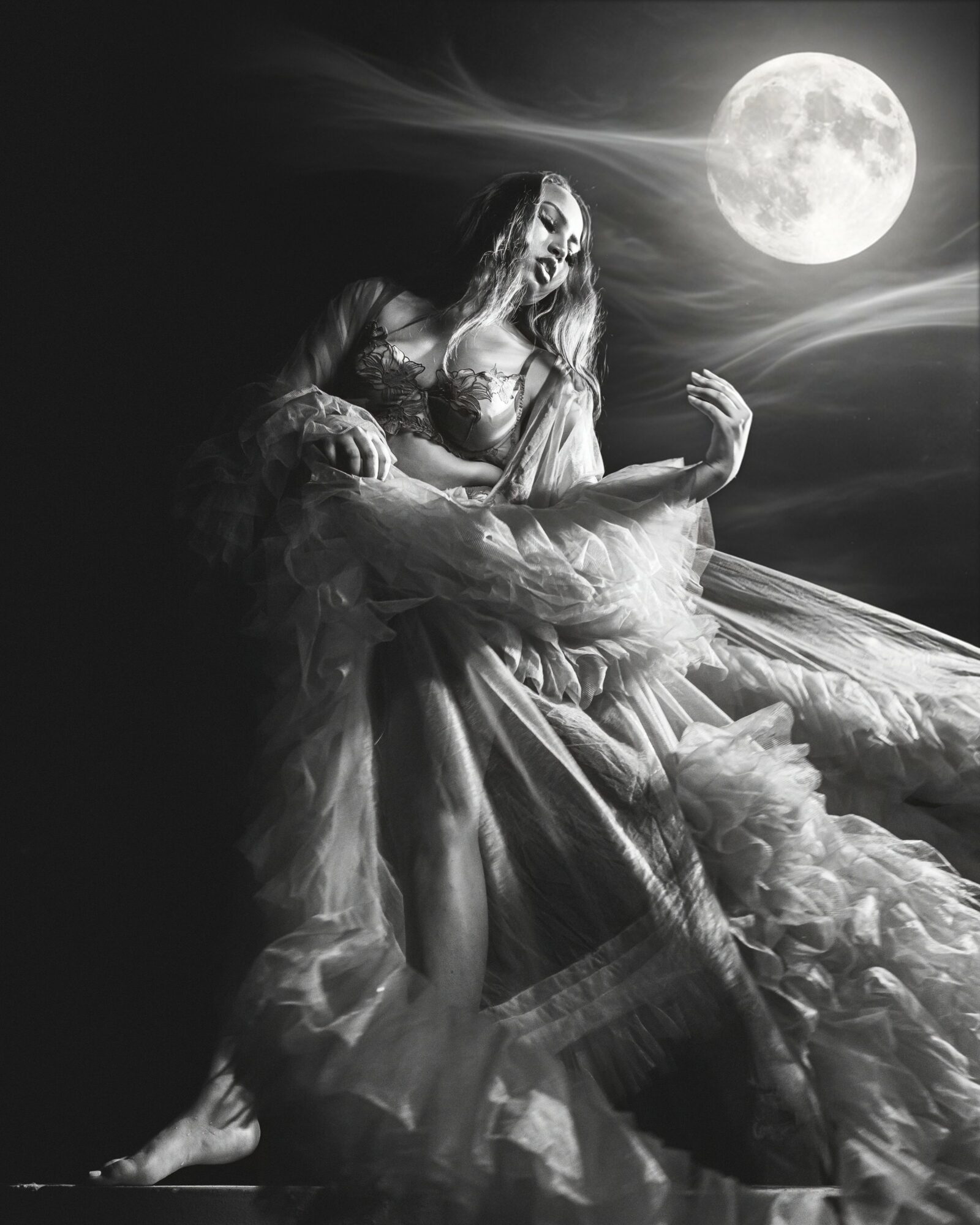 A woman in a flowing dress sits outdoors under a full moon, with clouds in the night sky, in black and white.