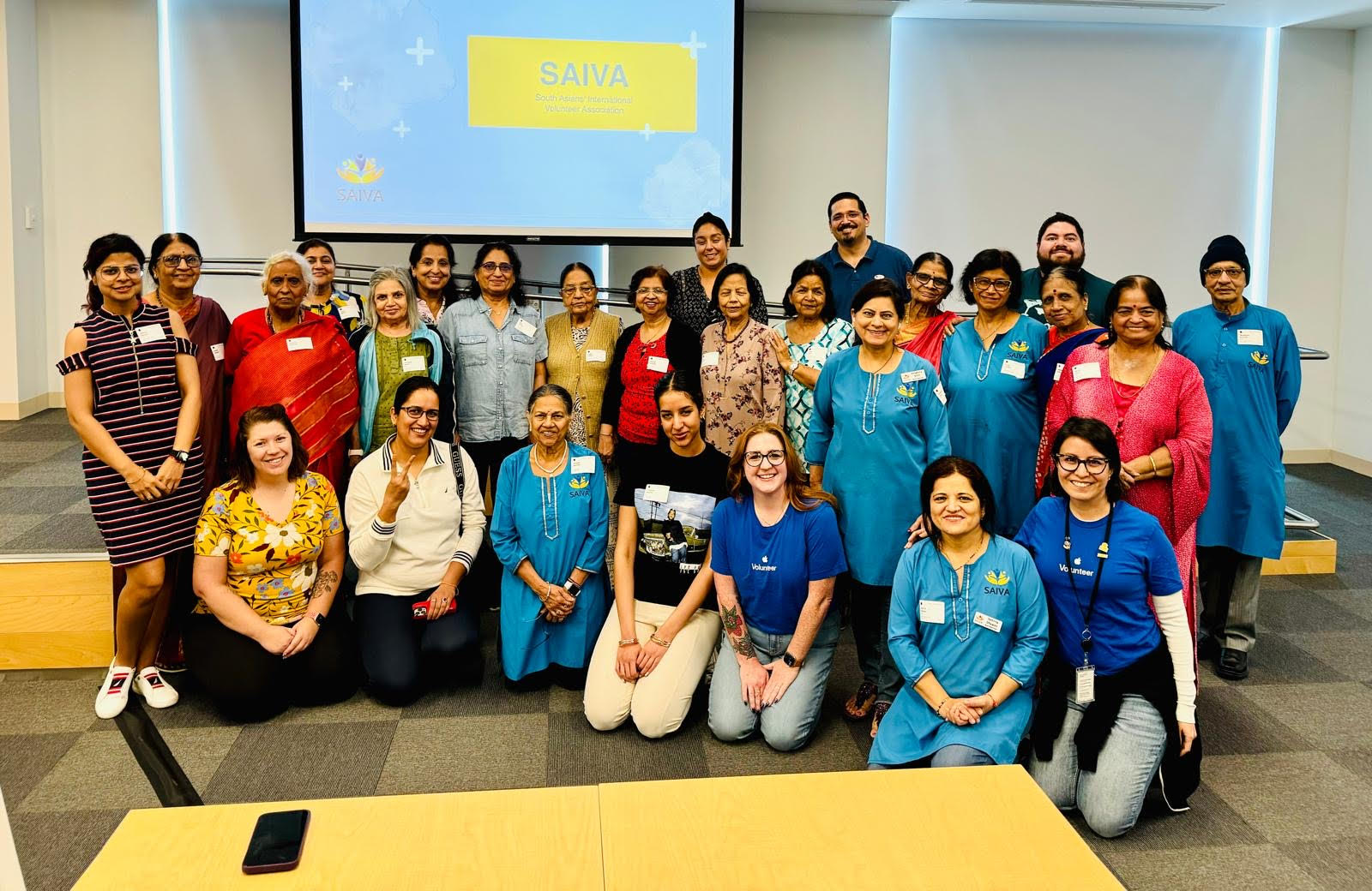 Group of 20 people in a room, some wearing blue shirts, standing and sitting in front of a screen displaying a yellow box with text. 20 words.
