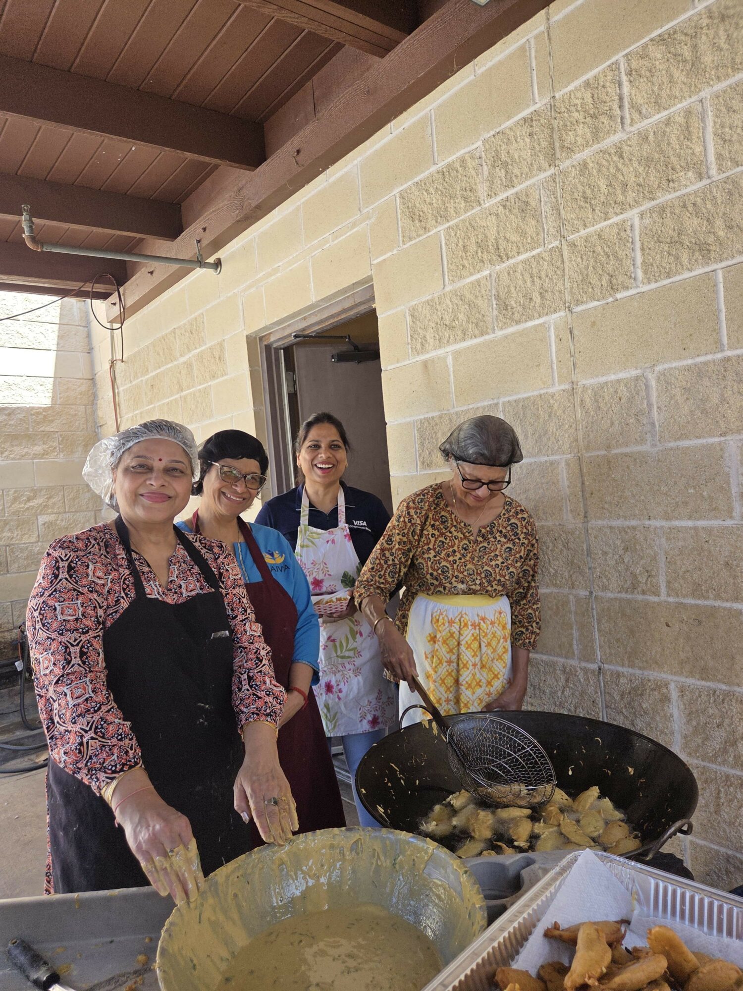 Four women cooking outdoors, wearing aprons and hairnets, standing near a large wok with food, against a brick wall.
