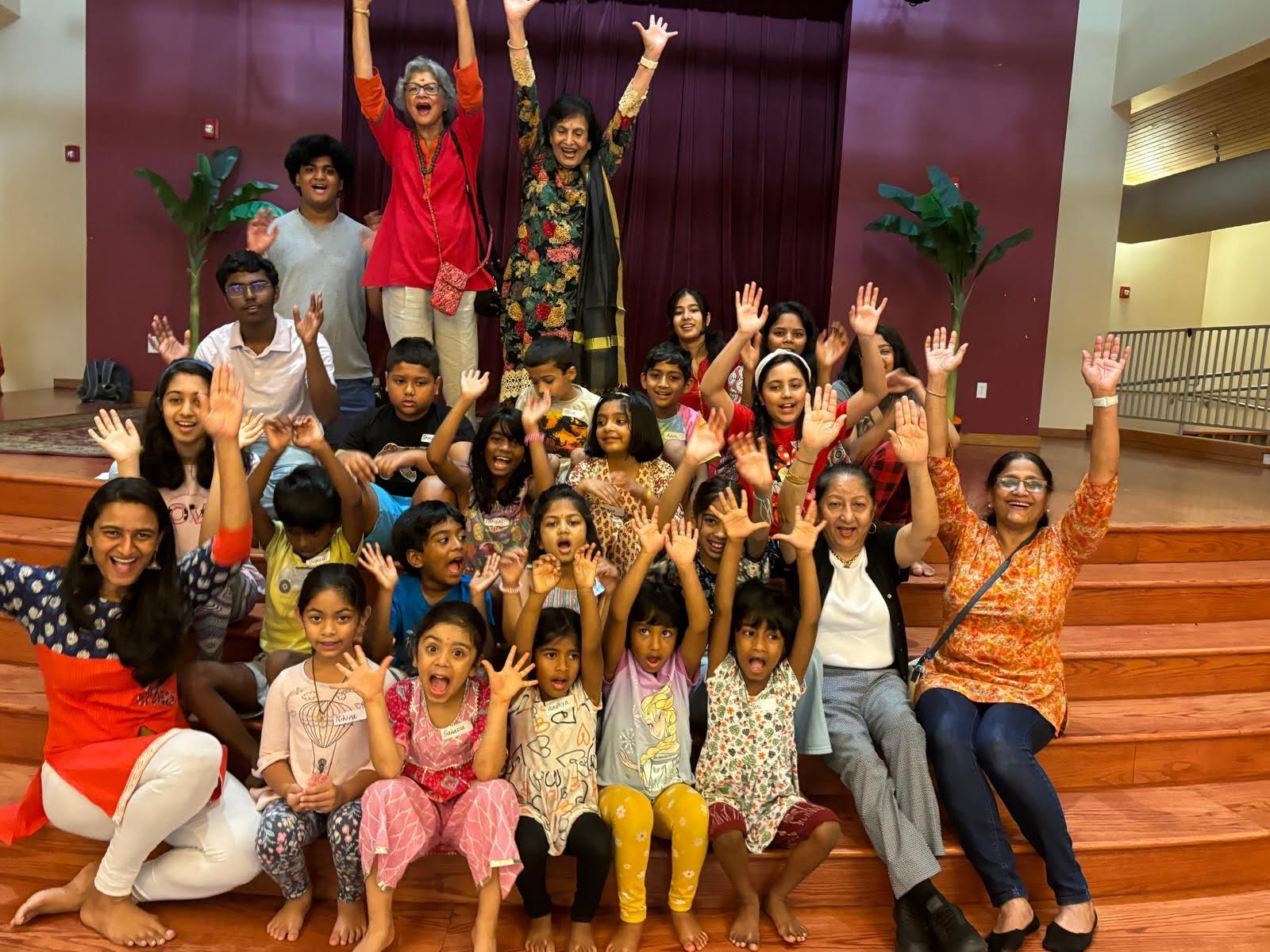 Group of children and adults on stage, smiling and waving, with purple curtains and potted plants in background.