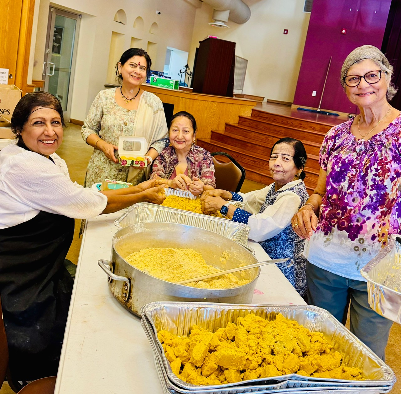 Four women and one girl gather around a table with large trays of yellow food, smiling and handing over a package.