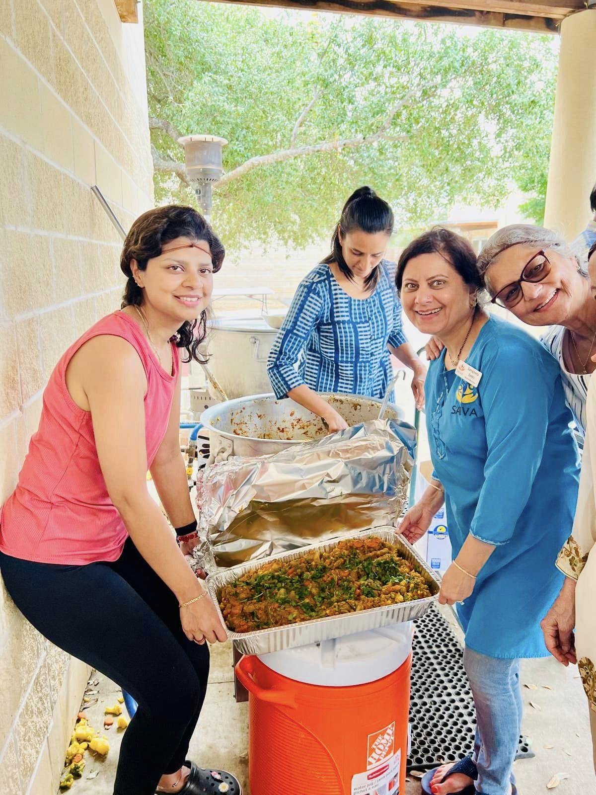 Four women and a girl are serving food outdoors, with greenery and trees in the background.