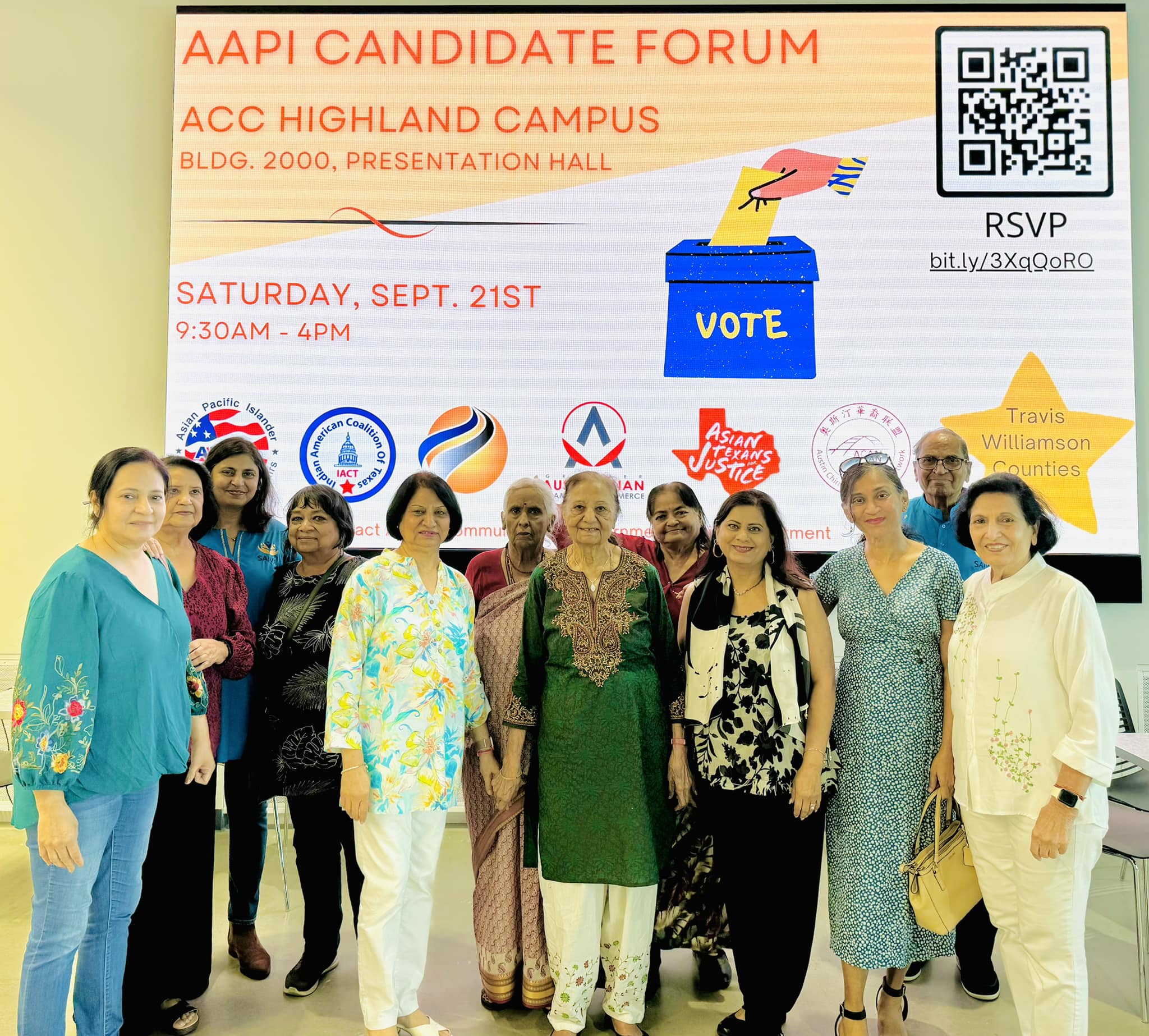 Group of nine people standing in front of a large event poster, smiling, indoors.