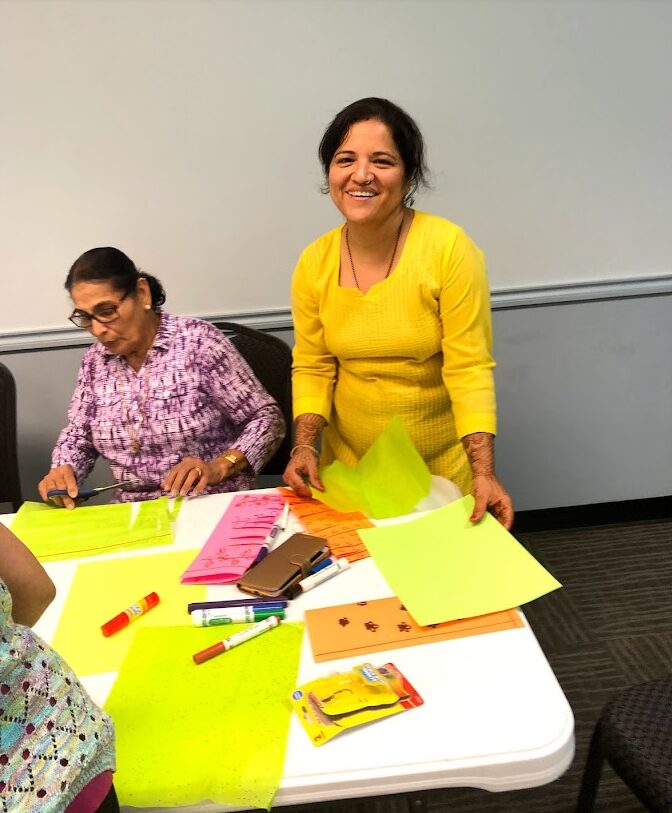 Two women at a table with colorful papers and supplies, one smiling and standing, the other sitting and working.