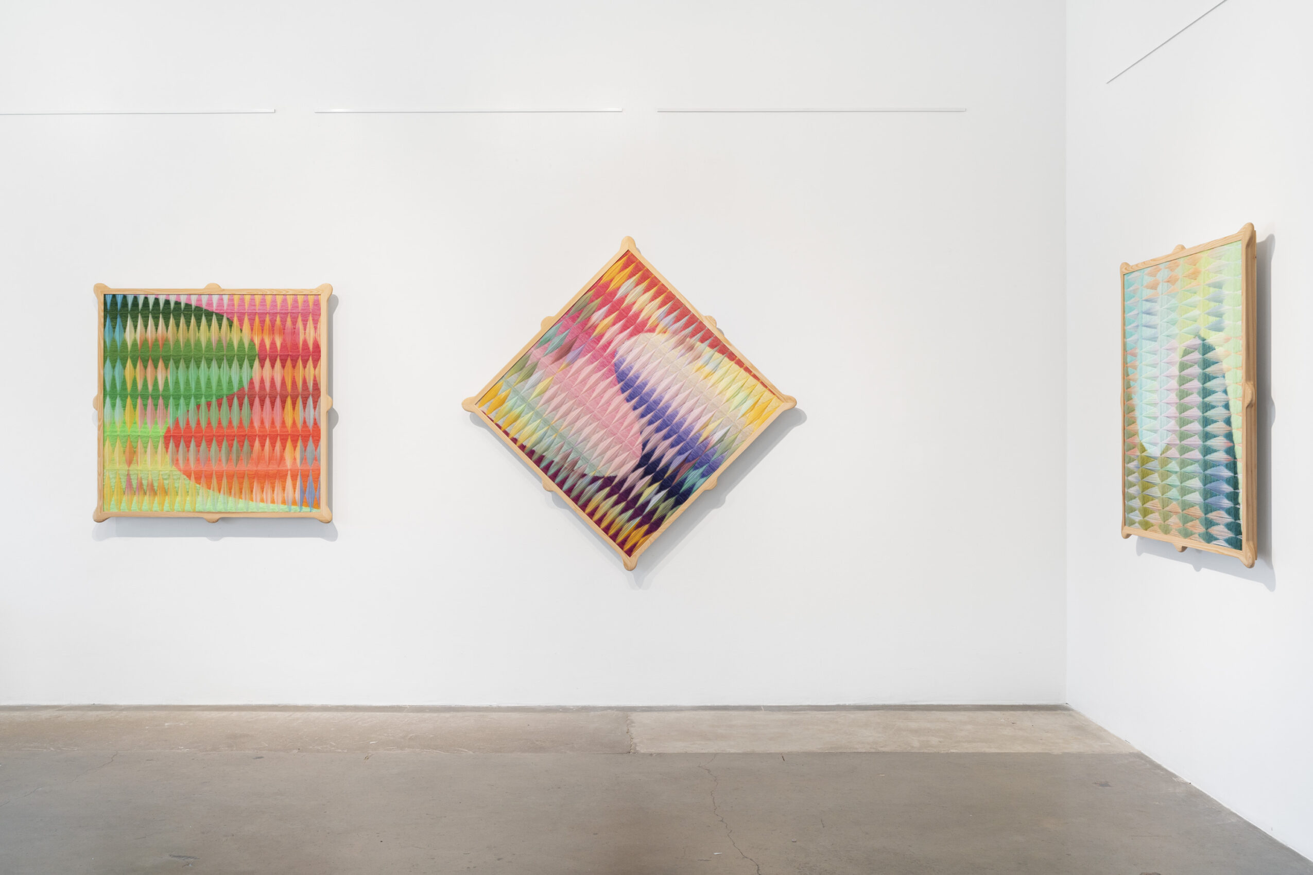 Three colorful abstract artworks on a white gallery wall, with a wooden floor below, arranged in a row with different orientations.