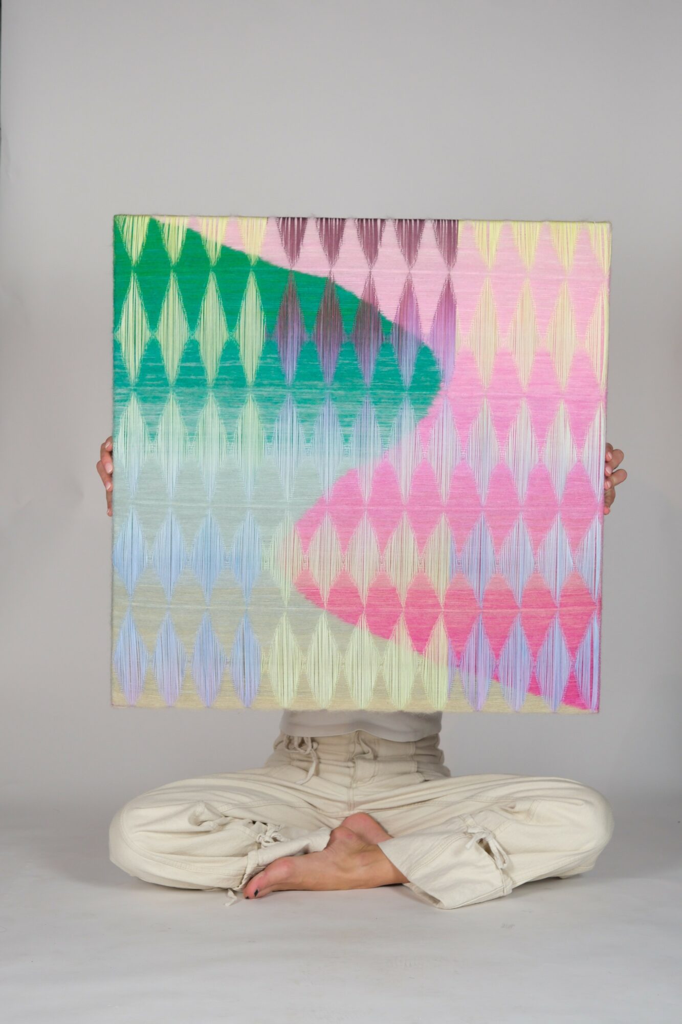 Person sitting cross-legged holding a colorful geometric patterned canvas in front of their face, against a plain background.