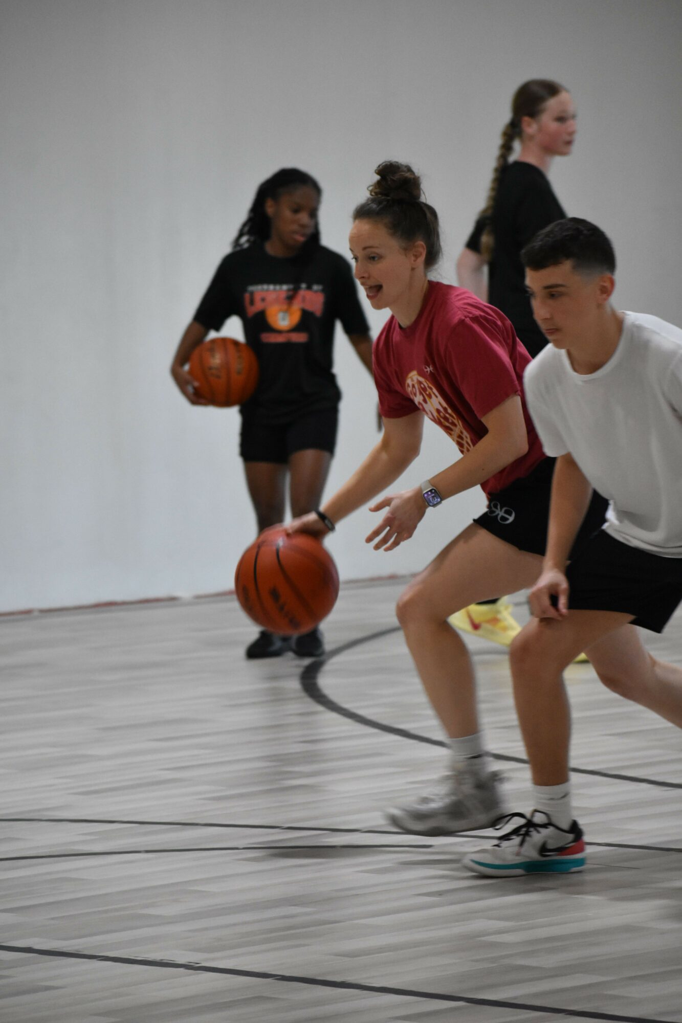 Two women playing basketball indoors, one in a pink shirt and black shorts, the other in a black shirt and shorts, with a coach nearby.