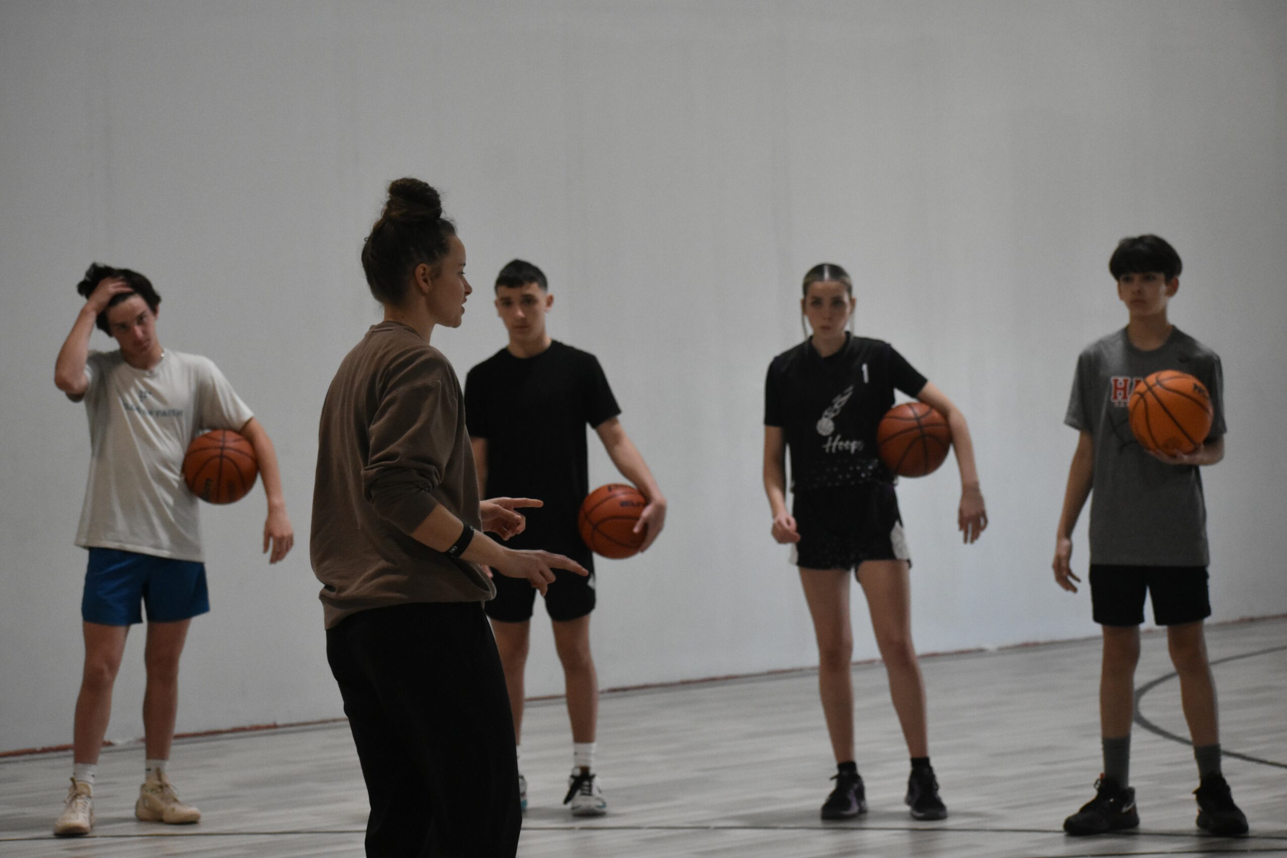 Four children holding basketballs in a gym, one girl in brown shirt, three boys in black and gray shirts, plain white wall background.