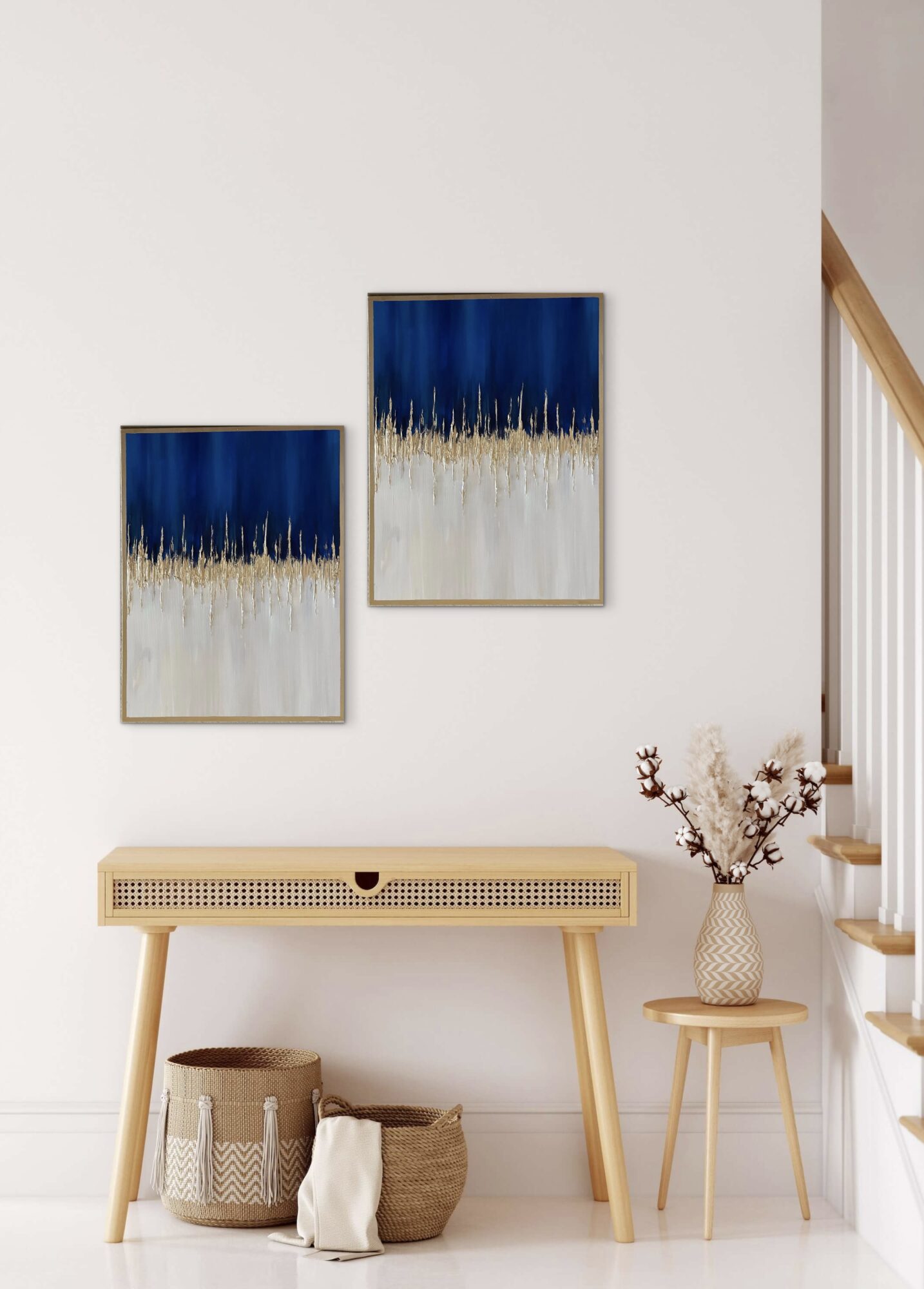 Two abstract paintings with blue, white, and gold colors hang above a wooden console table with baskets, next to a small table with a vase of flowers.