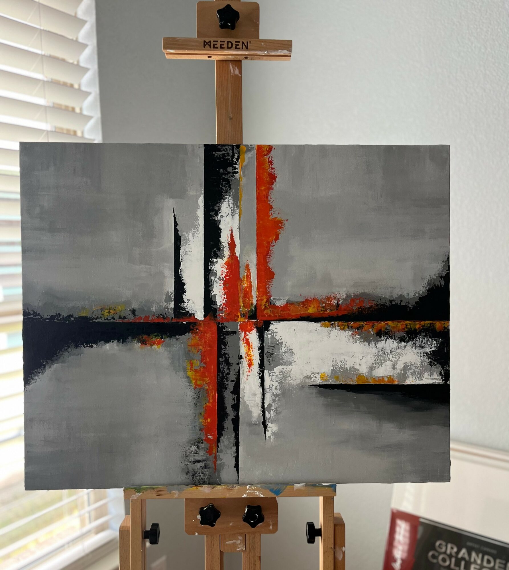 Abstract painting with vertical and horizontal lines in gray, black, white, and orange colors on a wall, with an easel and window nearby.
