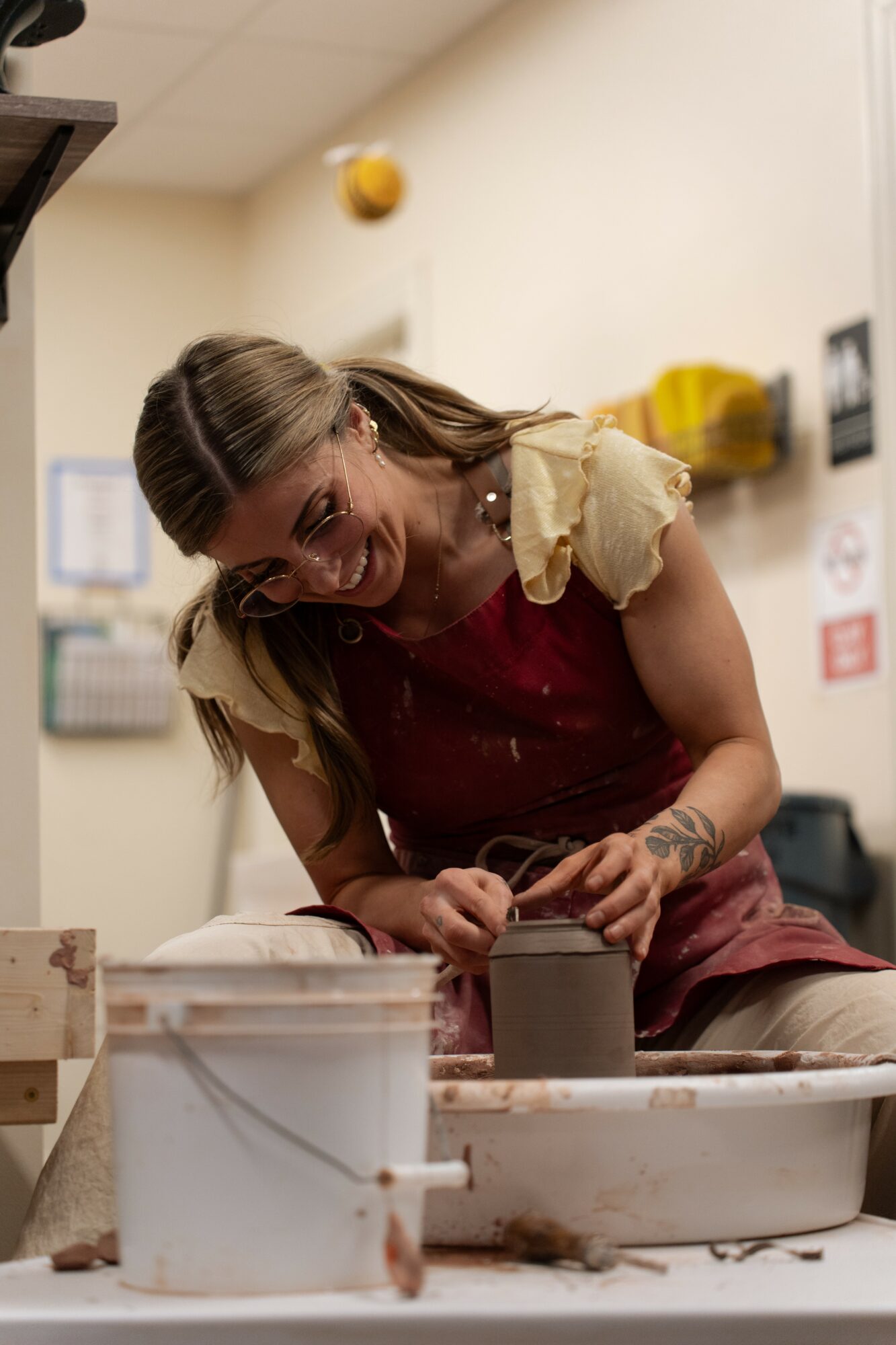Young woman with long hair smiling, leaning over a table, working on a project with various containers and tools.