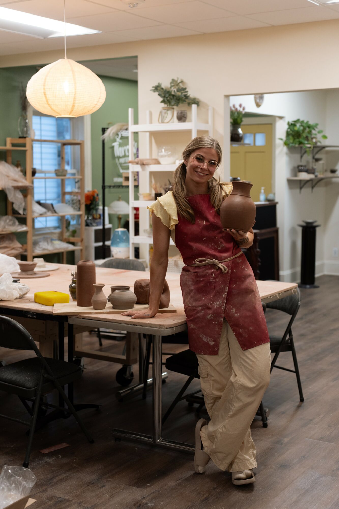 Woman in apron holding a pottery piece in a well-lit room with shelves and plants, standing near a table.