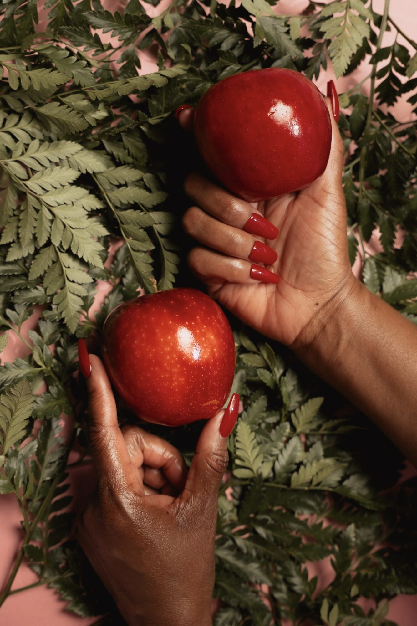 Two hands with red nail polish hold red apples among green fern leaves, background pink.