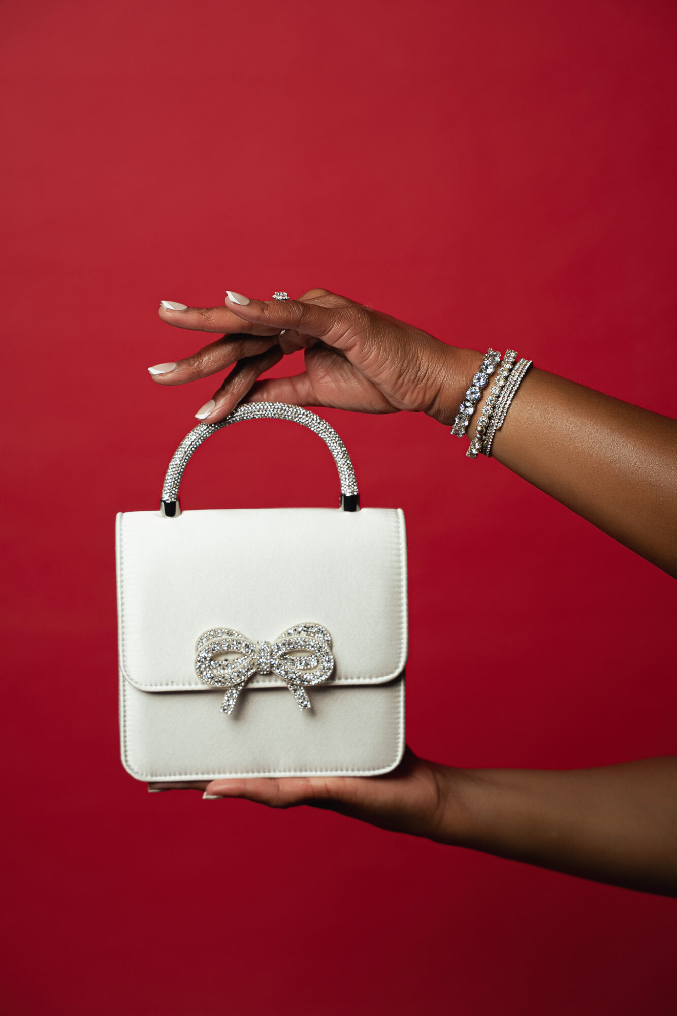 Person holding a white handbag with a bow, against a red background, wearing bracelets on wrist.