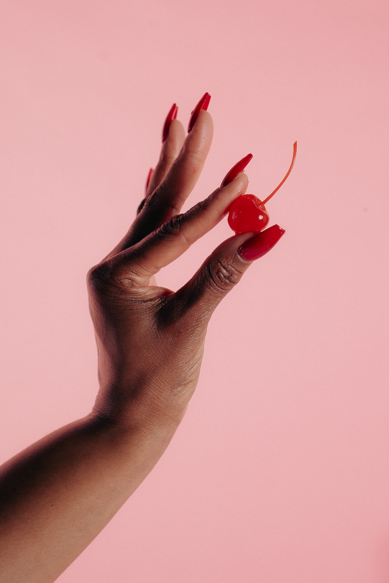 Hand with red nail polish holding a cherry against a pink background.