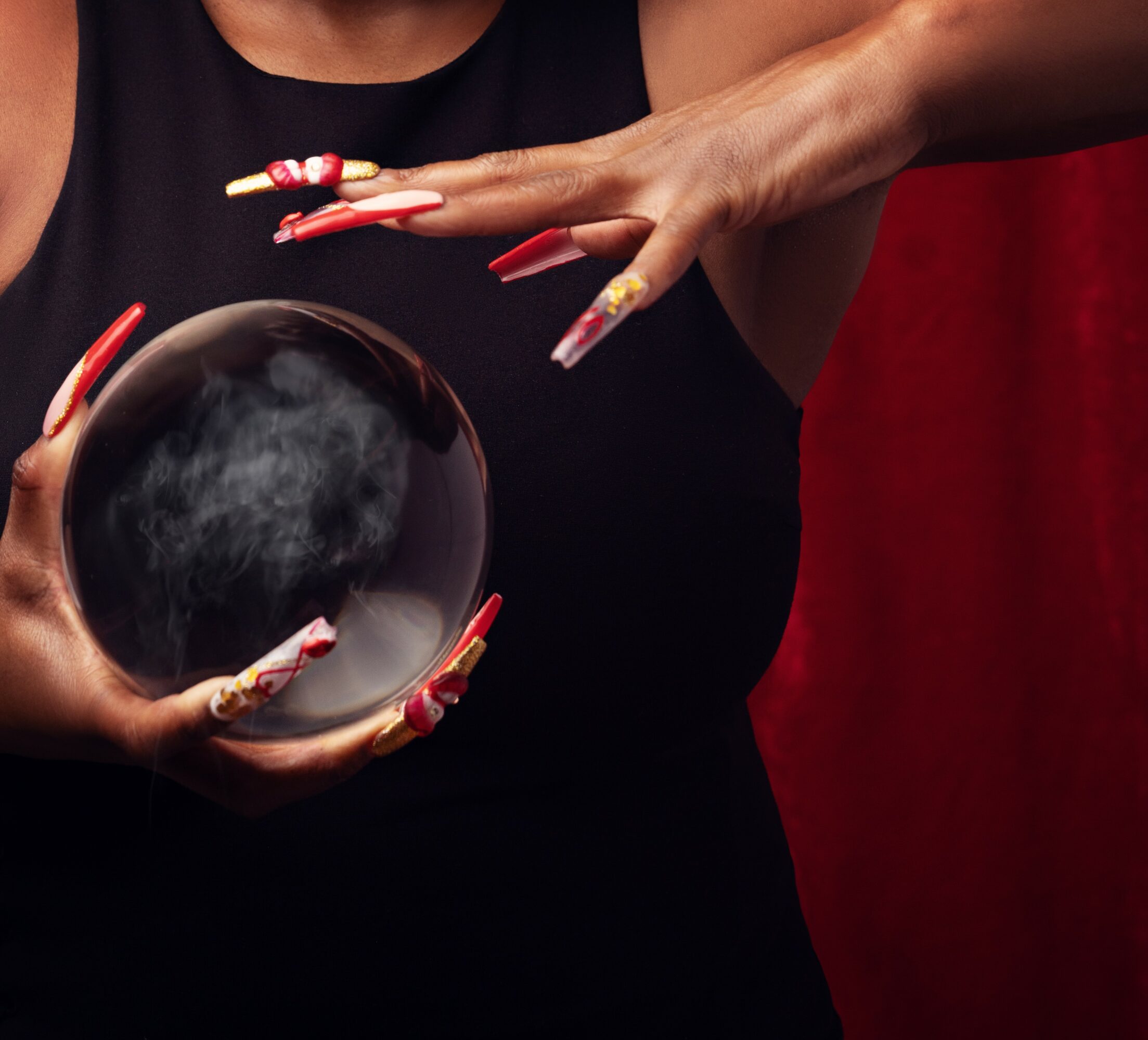 Person holding a crystal ball with long, decorated nails, wearing a black sleeveless top, against a red background.