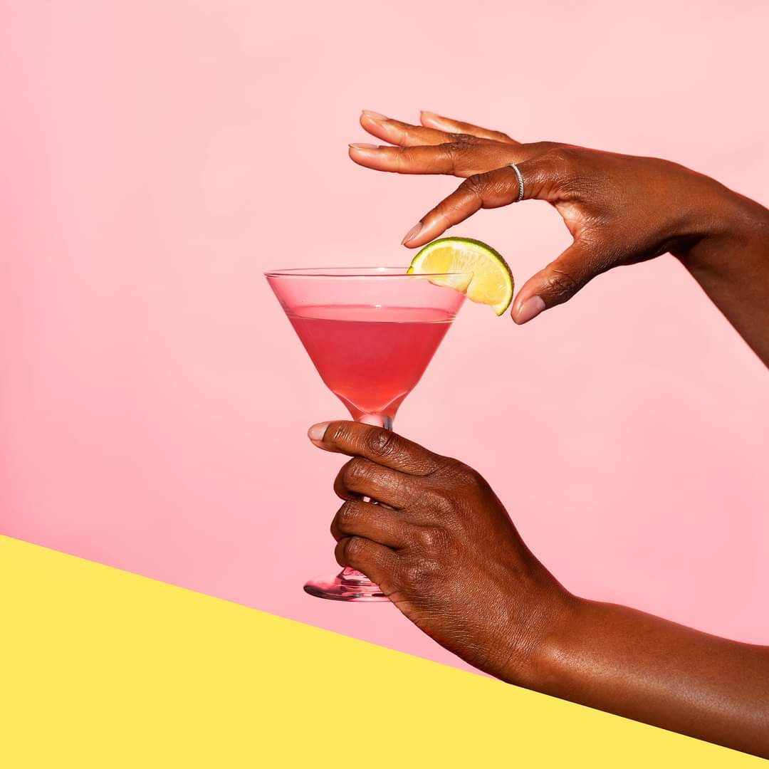 A hand holding a pink cocktail glass with a lemon wedge, against a pink and yellow background.
