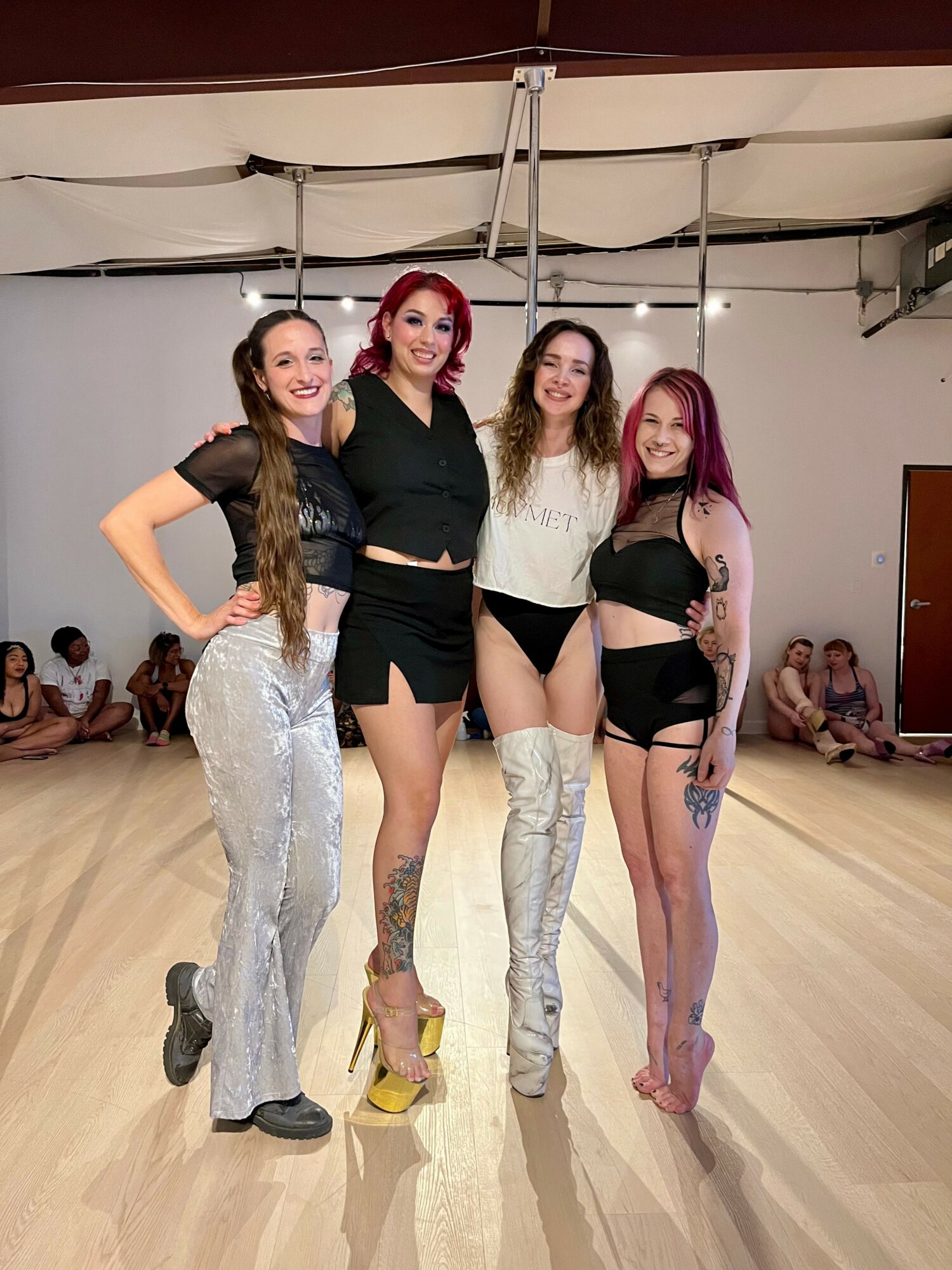 Four women stand together in a dance studio, smiling, with a group of people sitting on the floor in the background.