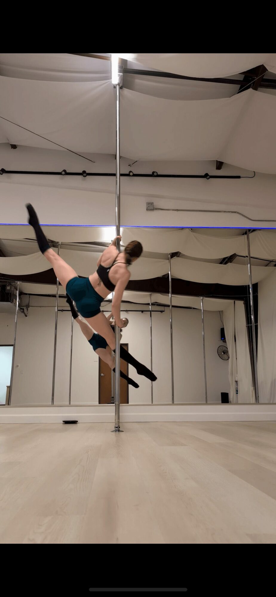 Person practicing pole dance in studio, holding pole with one hand and leg extended outward.