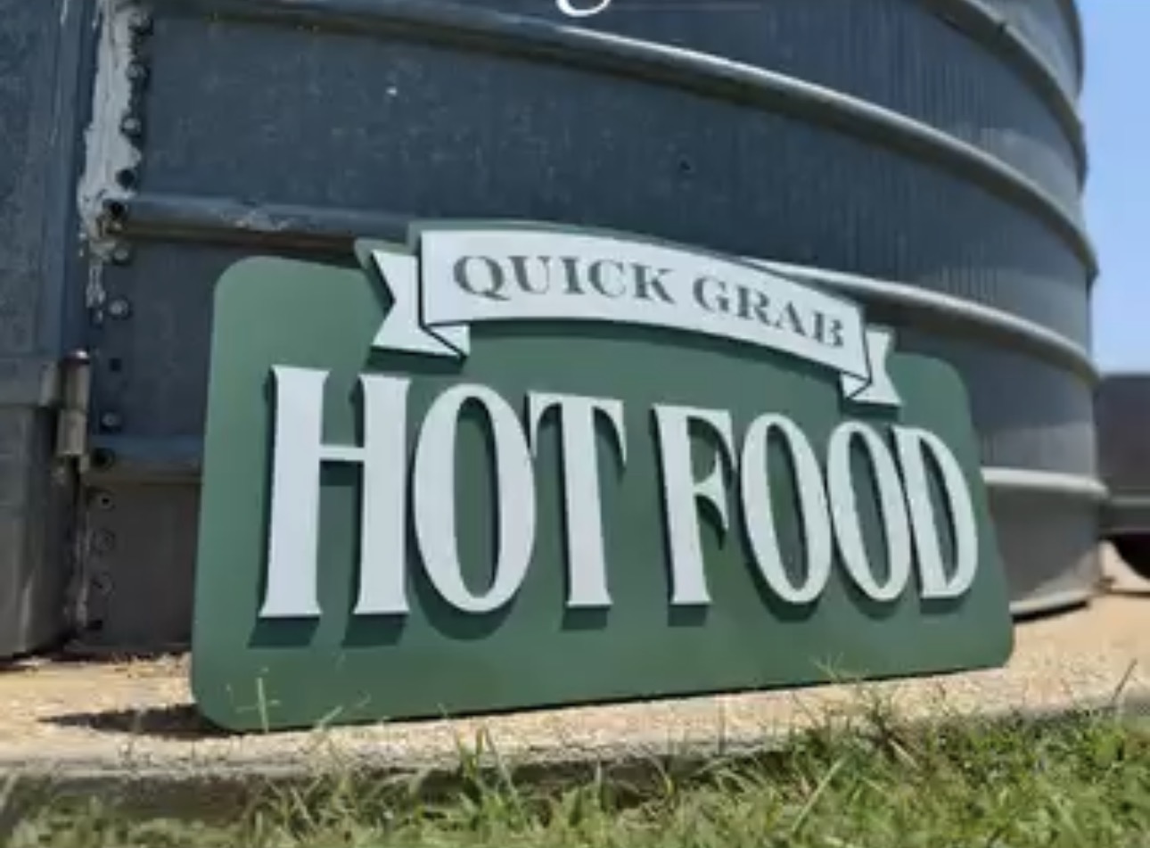 Sign advertising quick grab hot food, placed on the ground near large containers, with grass in the foreground.