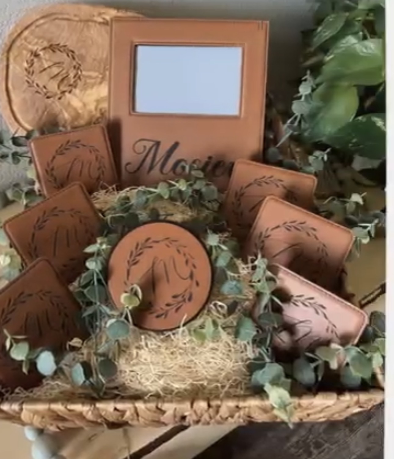 Decorative makeup mirror and makeup containers arranged in a basket with greenery, on a surface with plants nearby.
