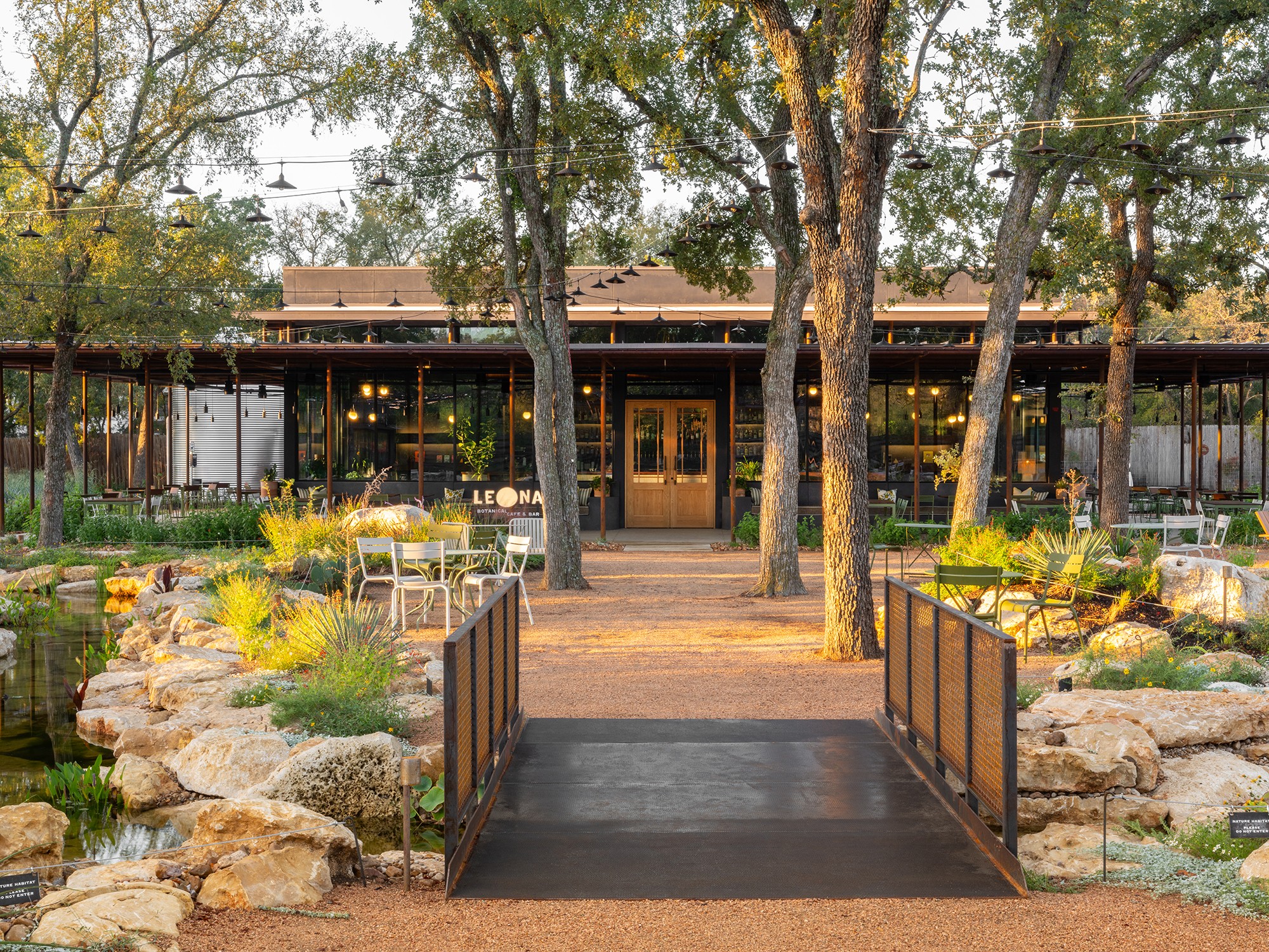 A building with a porch, trees, and a garden with rocks and plants in front, viewed from a bridge.