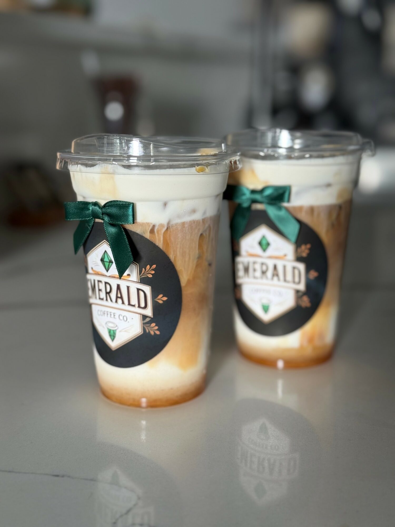 Two cups of layered coffee drinks with whipped cream, decorated with green ribbons, on a reflective surface.