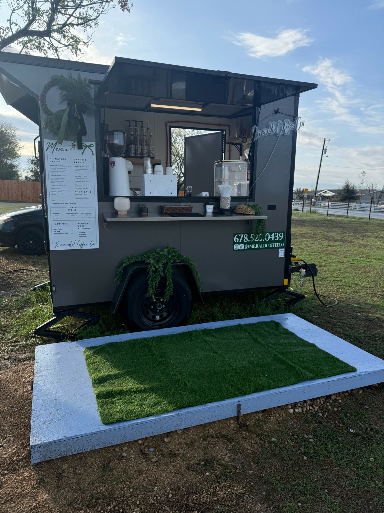 Food truck with a menu, serving window, and a small patch of artificial grass outside, under a partly cloudy sky.