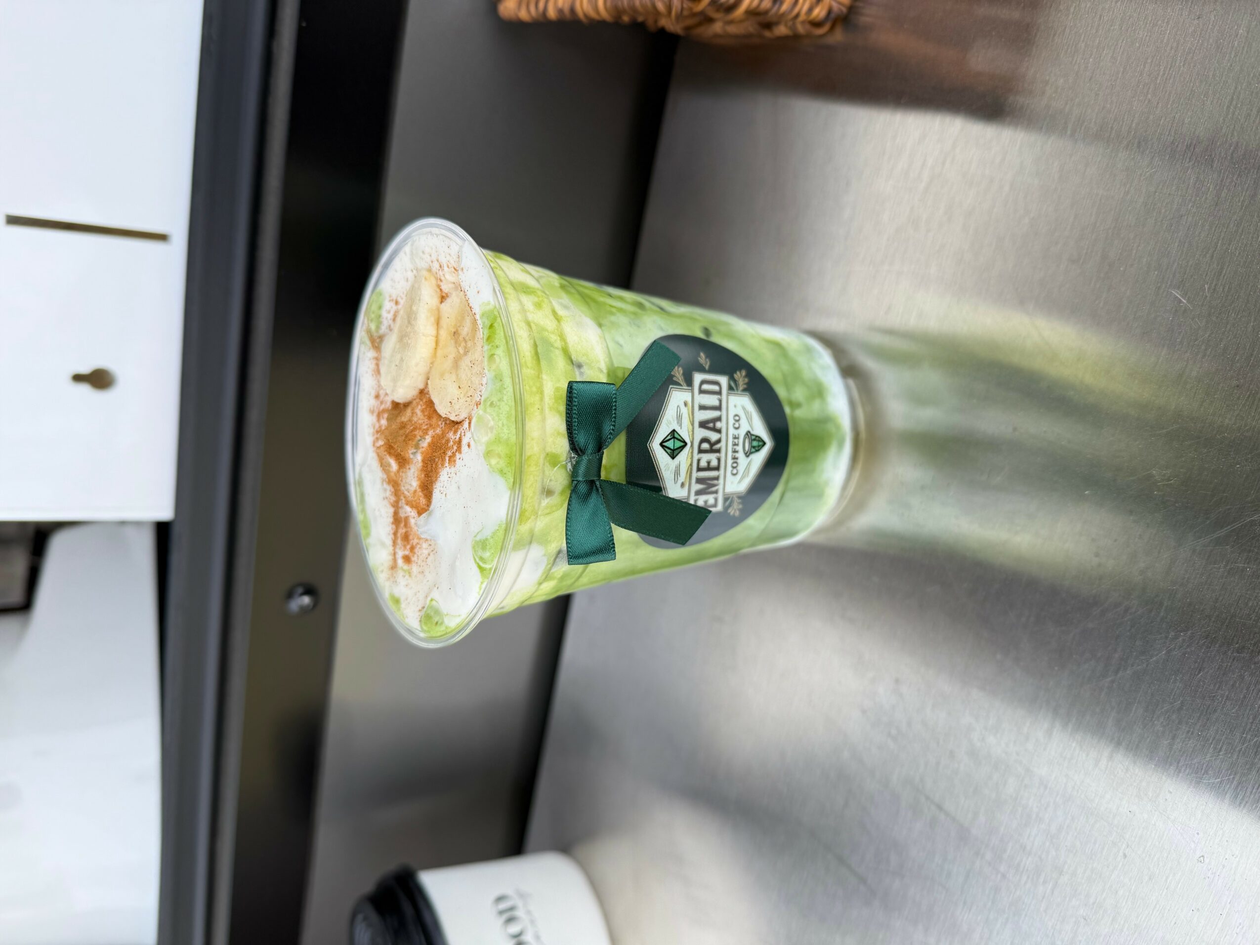 Iced beverage in a clear cup with a green straw and a green ribbon, on a metallic surface.