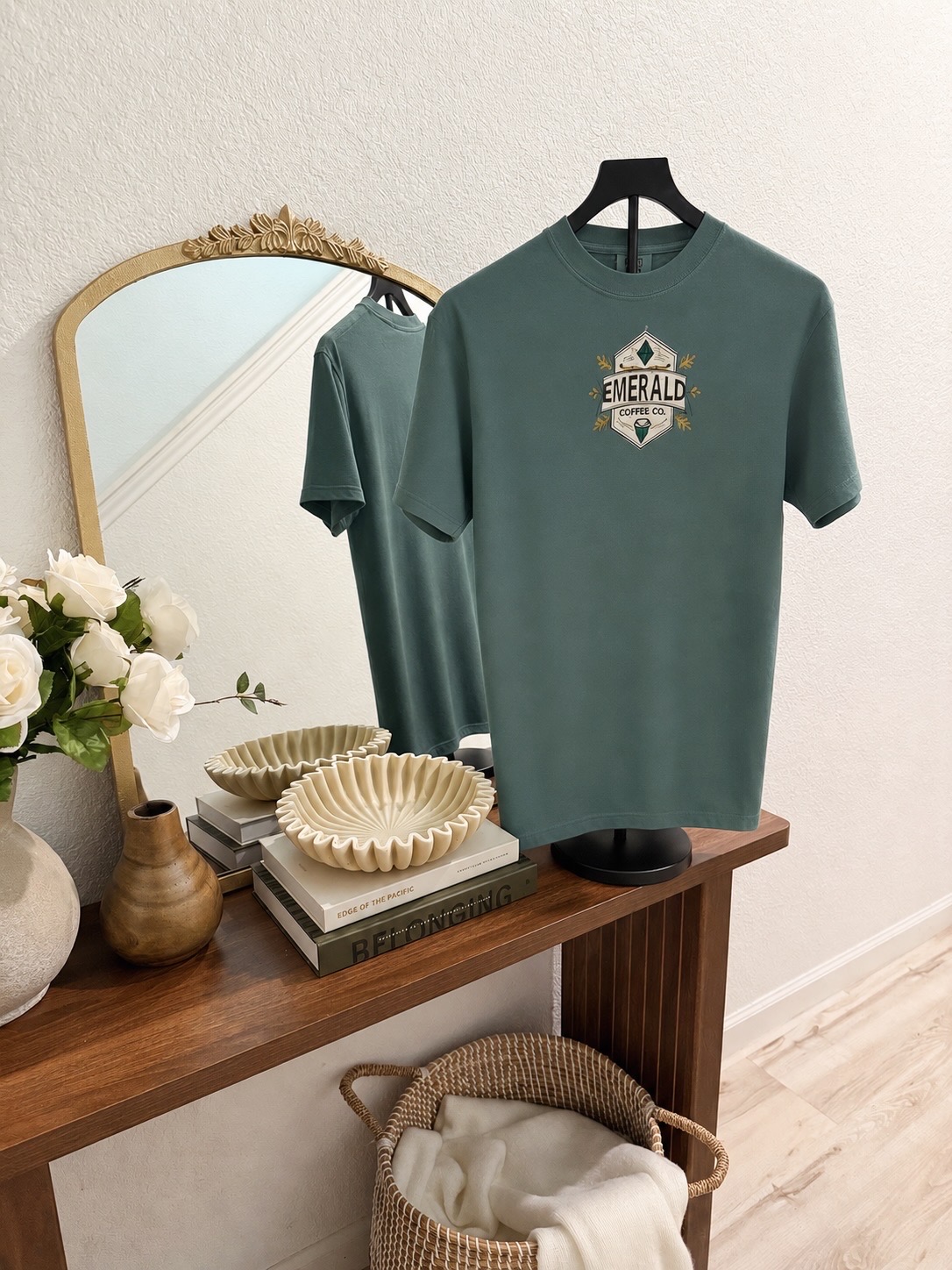 Teal T-shirt with a logo hangs on a black hanger, reflected in a mirror above a wooden table with flowers and bowls.