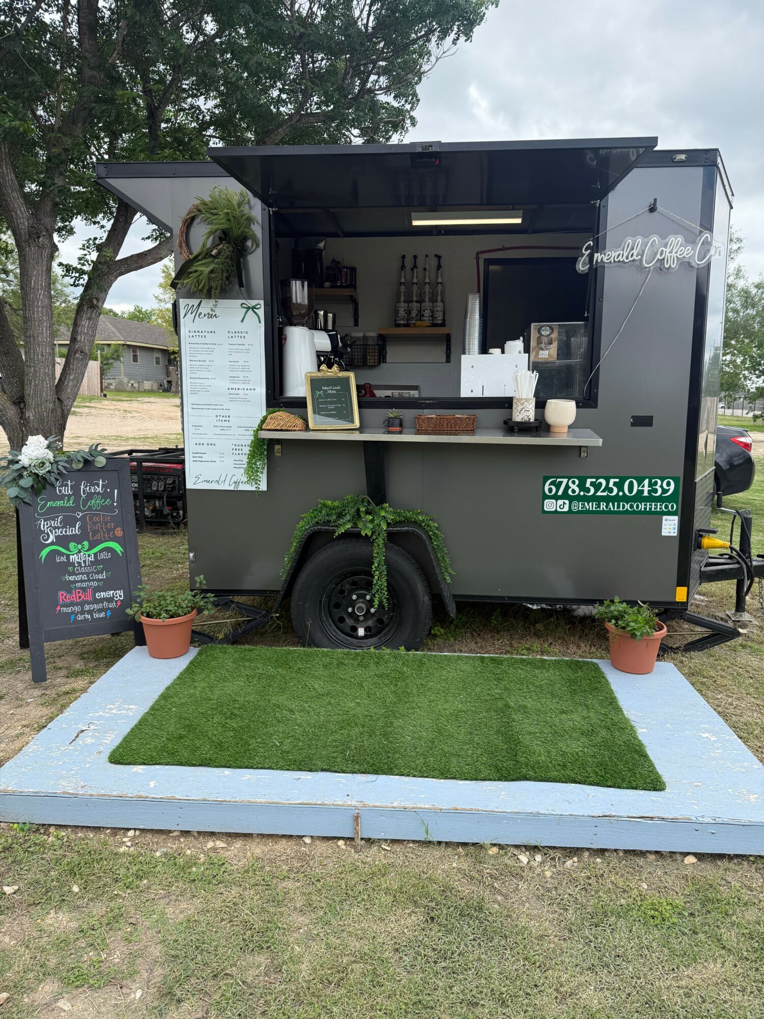 Food truck with menu board, black tires, and small green artificial grass area, parked on grass with trees in background.