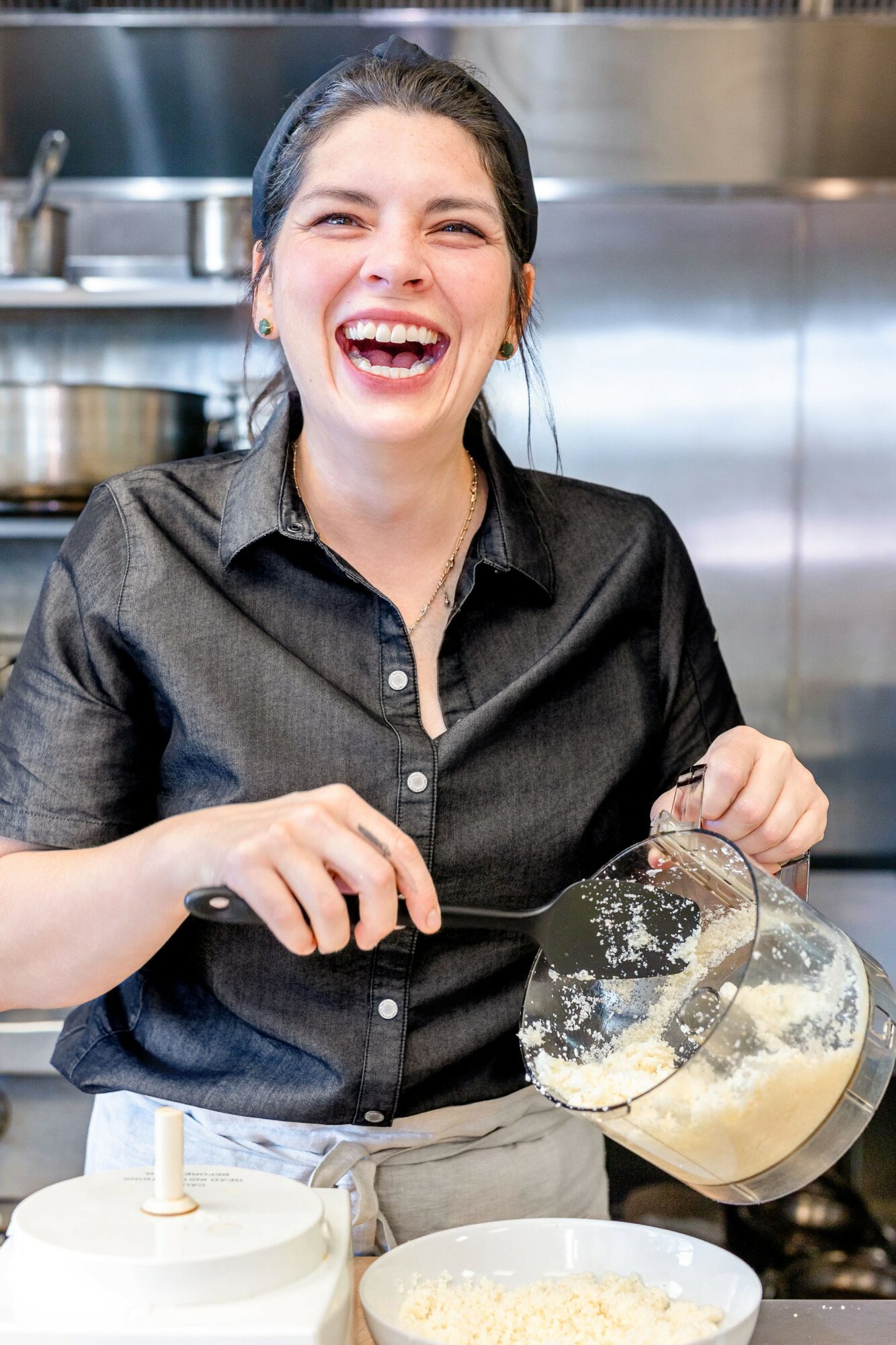 Woman laughing while pouring batter into a bowl in a kitchen, wearing a dark shirt.
