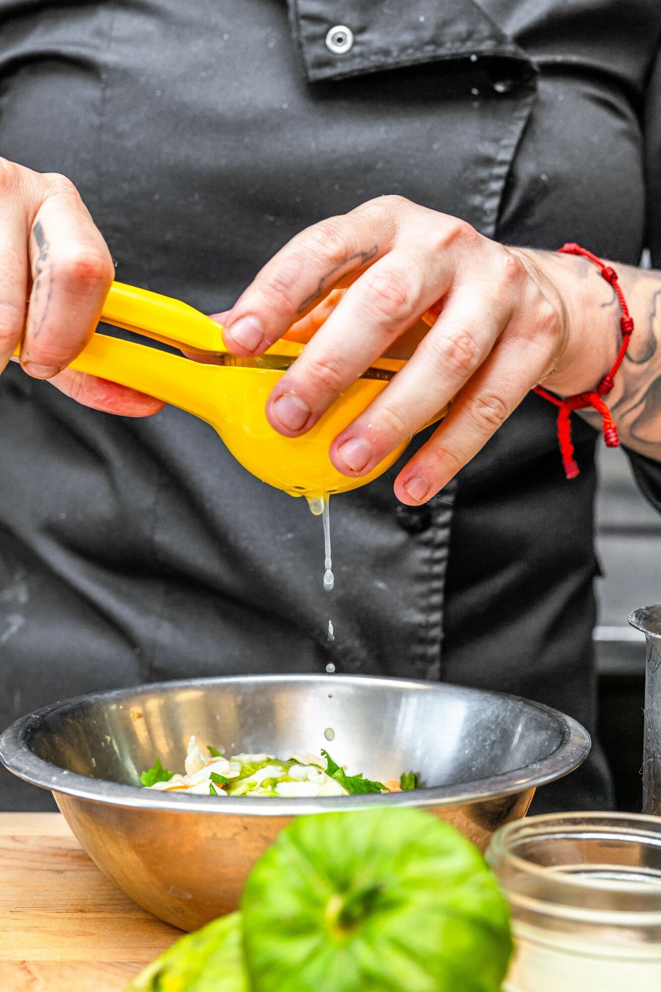 Person grating a yellow vegetable peeler over a metal bowl with sliced vegetables, on a wooden surface, with a green vegetable in foreground.