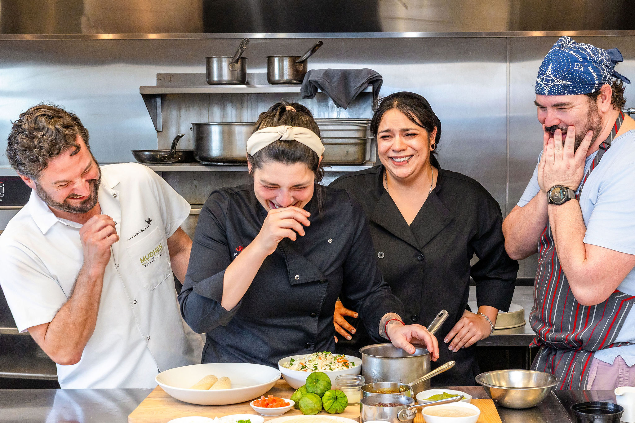Four chefs in a kitchen laughing and sharing a moment, with bowls and vegetables on the counter.