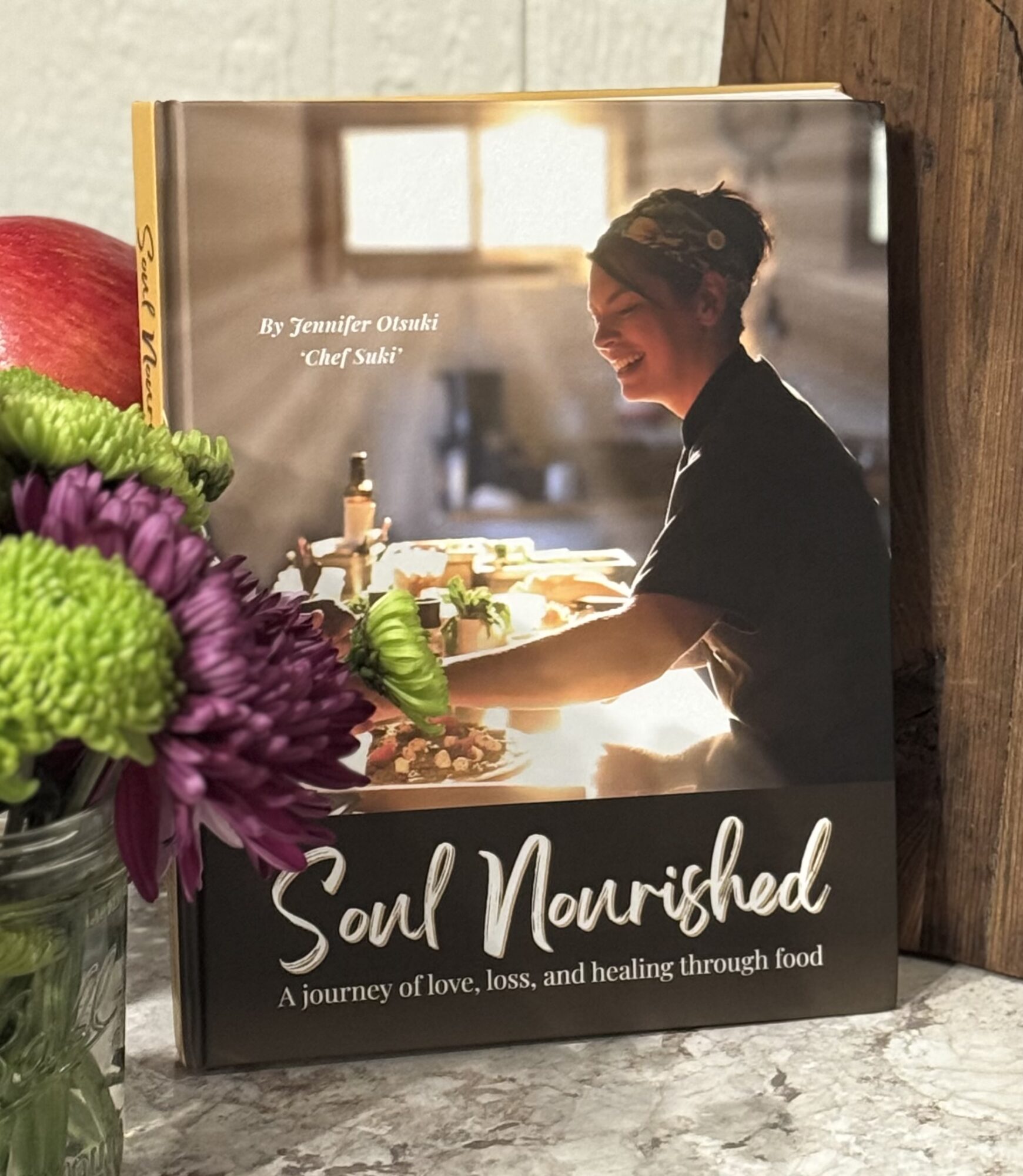 Book cover featuring a smiling woman in a kitchen, with sunlight streaming through a window, and a bouquet of flowers nearby.