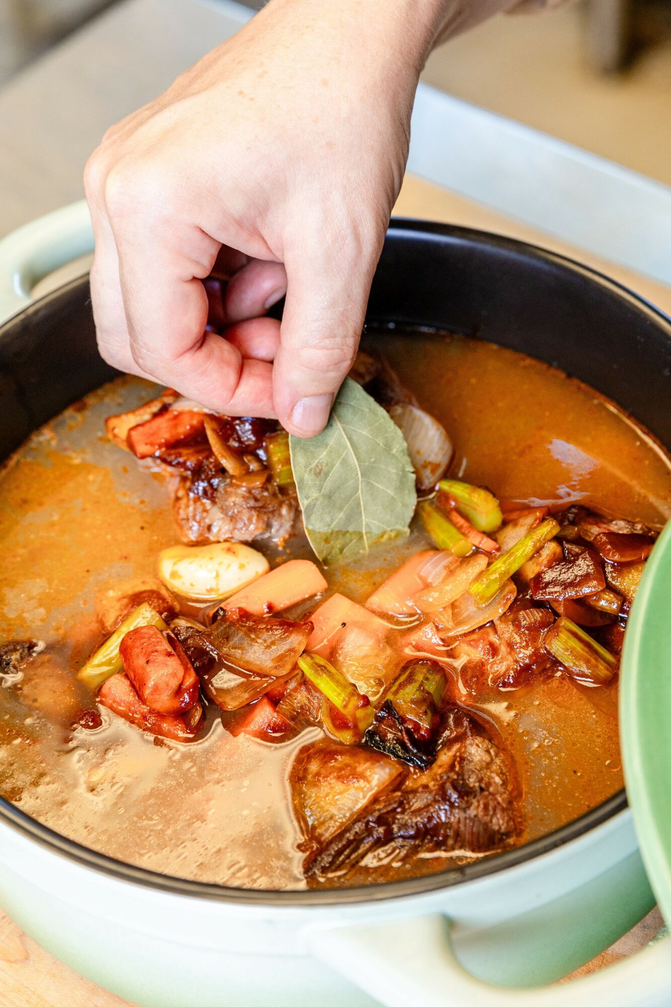 Hand adding a bay leaf to a pot of stew with meat, vegetables, and broth, on a stovetop.