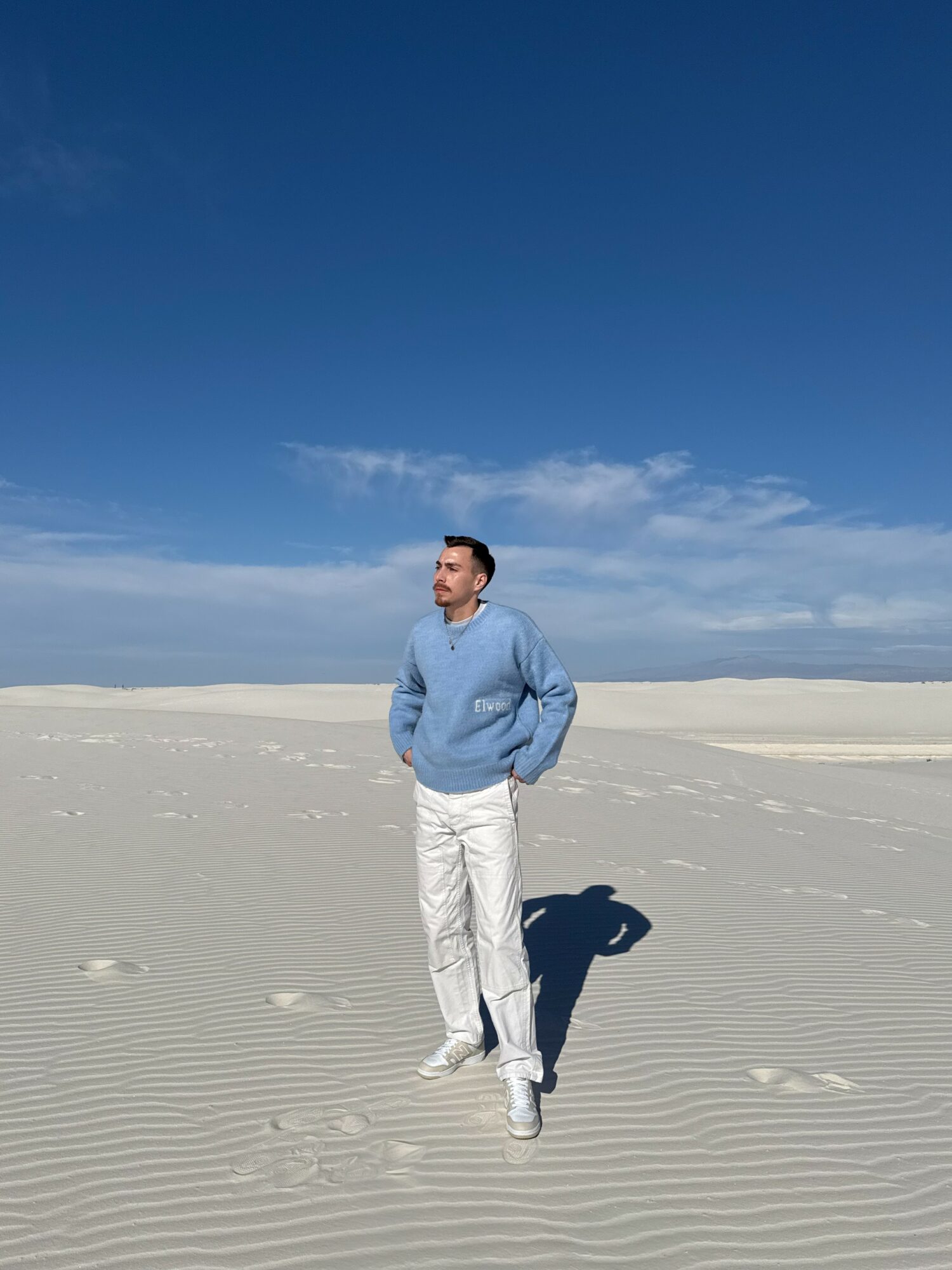 Man standing on sandy beach with blue sky and clouds, wearing a blue sweater and white pants, shadow visible.