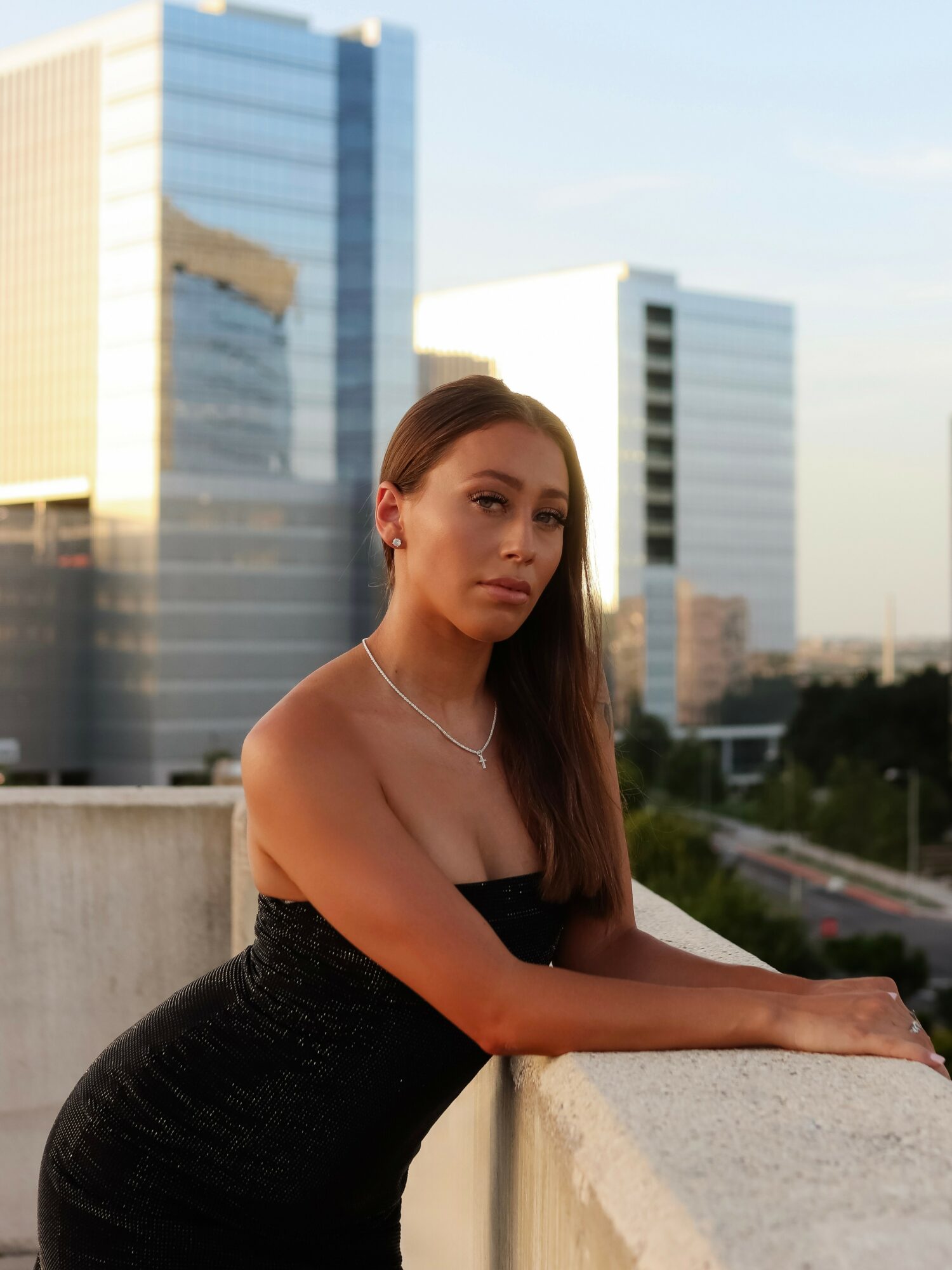 Young woman with long hair in a strapless black dress leaning on a concrete ledge with city buildings in the background.